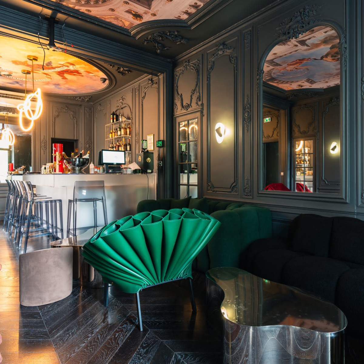 A stylish lounge area featuring a bar with a modern design, ornate wall decor, and plush seating arrangements in various colors.