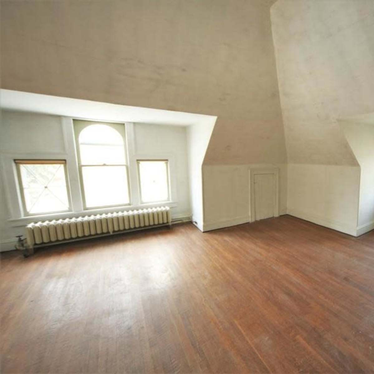 A spacious, empty room with sloped walls, wooden floors, and two windows allowing light to enter.