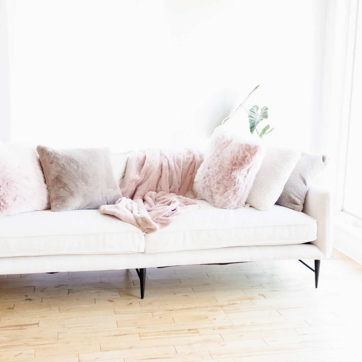 A light-colored sofa with plush pillows and a pink throw blanket is positioned near large windows on a wooden floor.