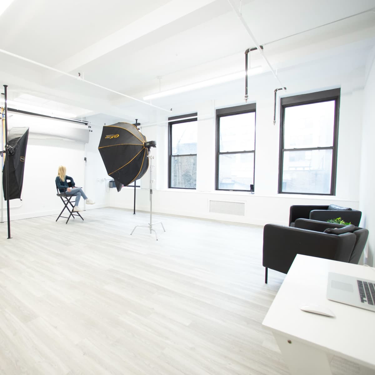 A bright photography studio with two large softboxes, a chair, and a small black sofa, all arranged in a minimally decorated space.