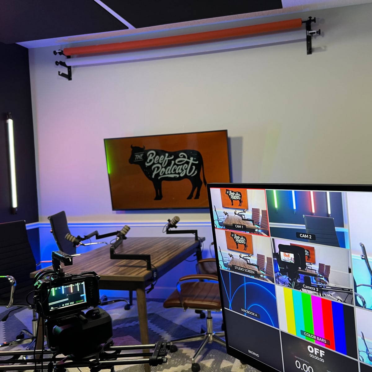 A podcast studio setup featuring a large screen displaying "The Beef Podcast," with multiple camera feeds visible on a monitor and microphones arranged on a table.