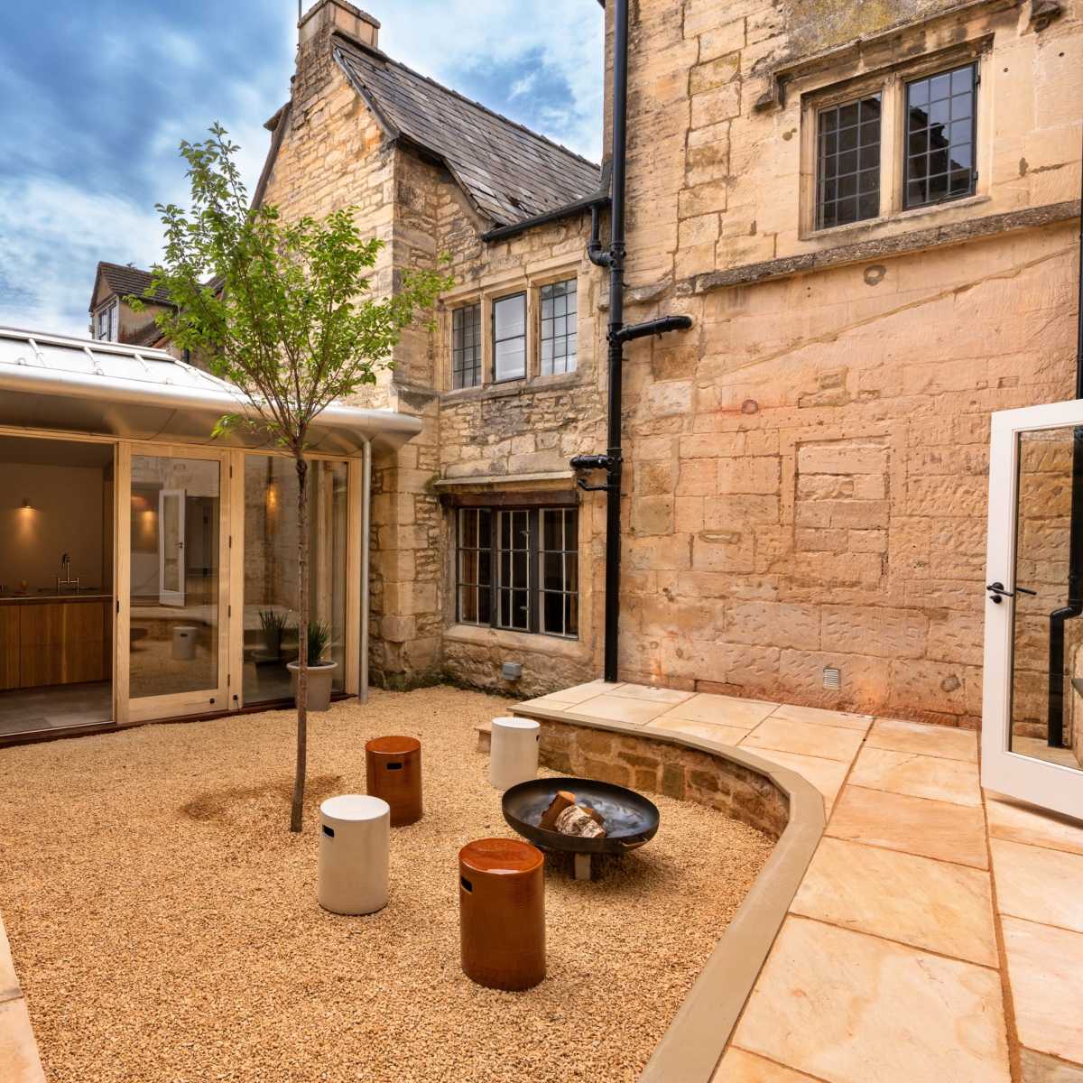 A stone courtyard surrounded by buildings, featuring a central tree, seating elements, and a fire pit.