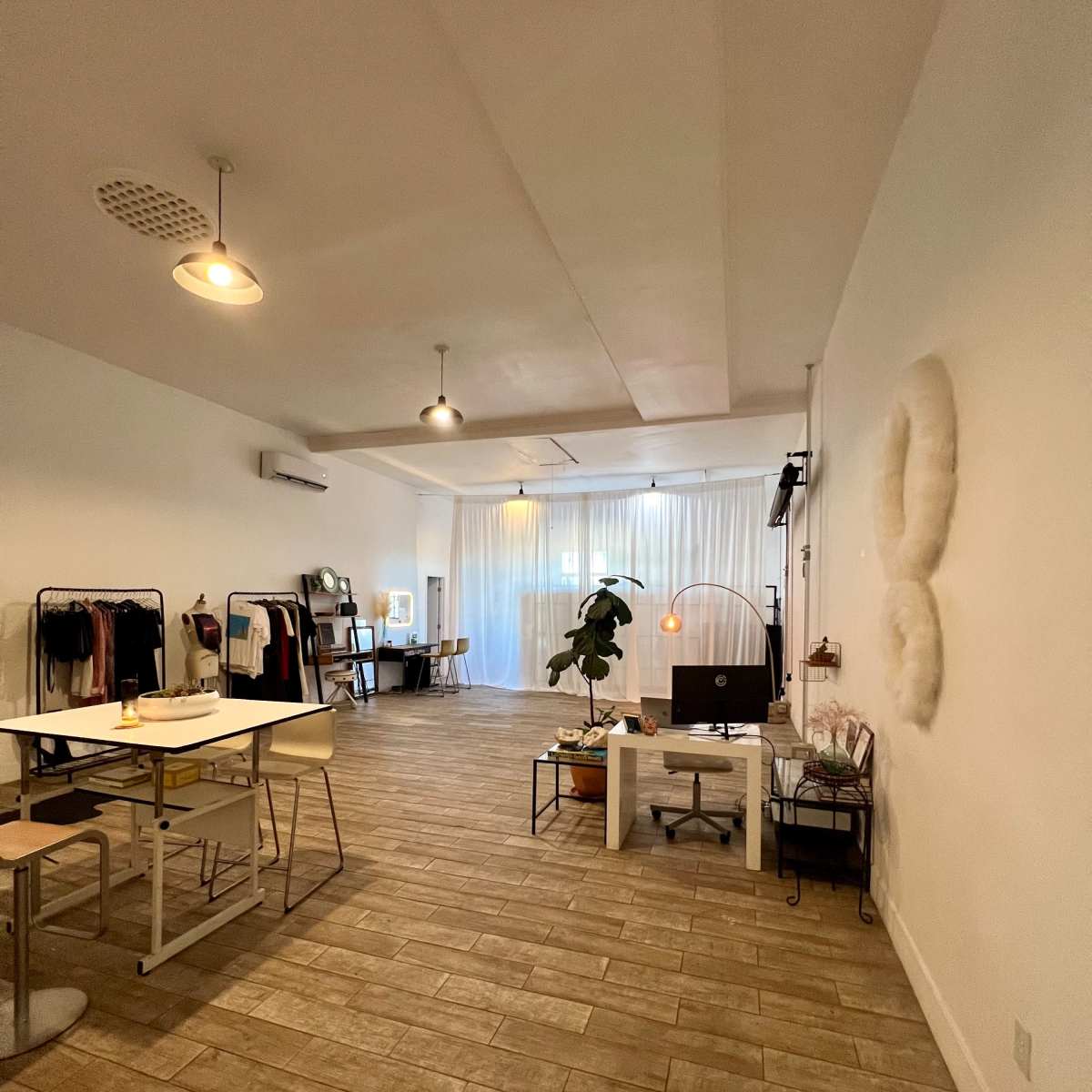 A spacious, minimalist room with a wooden floor, clothing racks, a small table with chairs, and light curtains letting in natural light.