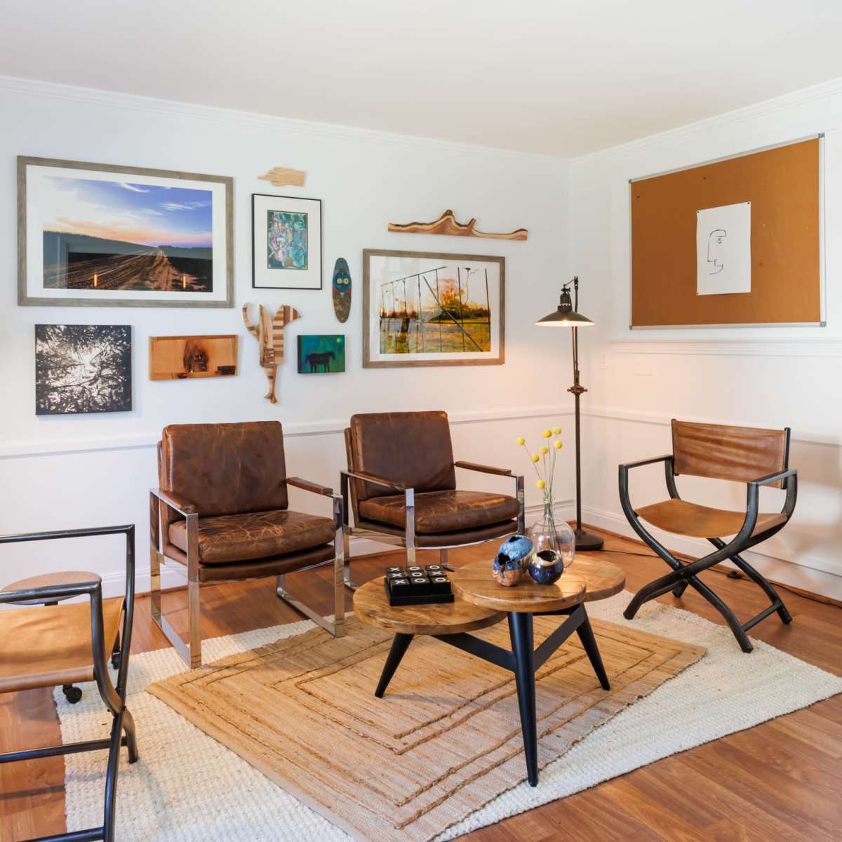 A cozy living room with four chairs arranged around a round coffee table on a woven rug, surrounded by wall art and a bookshelf filled with decorative items.