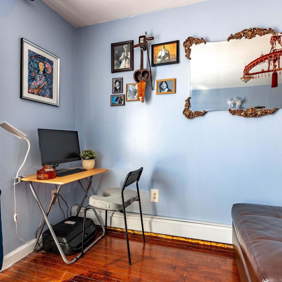A small room features a desk with a computer, a leather couch, and a wall decorated with framed pictures and a large mirror.