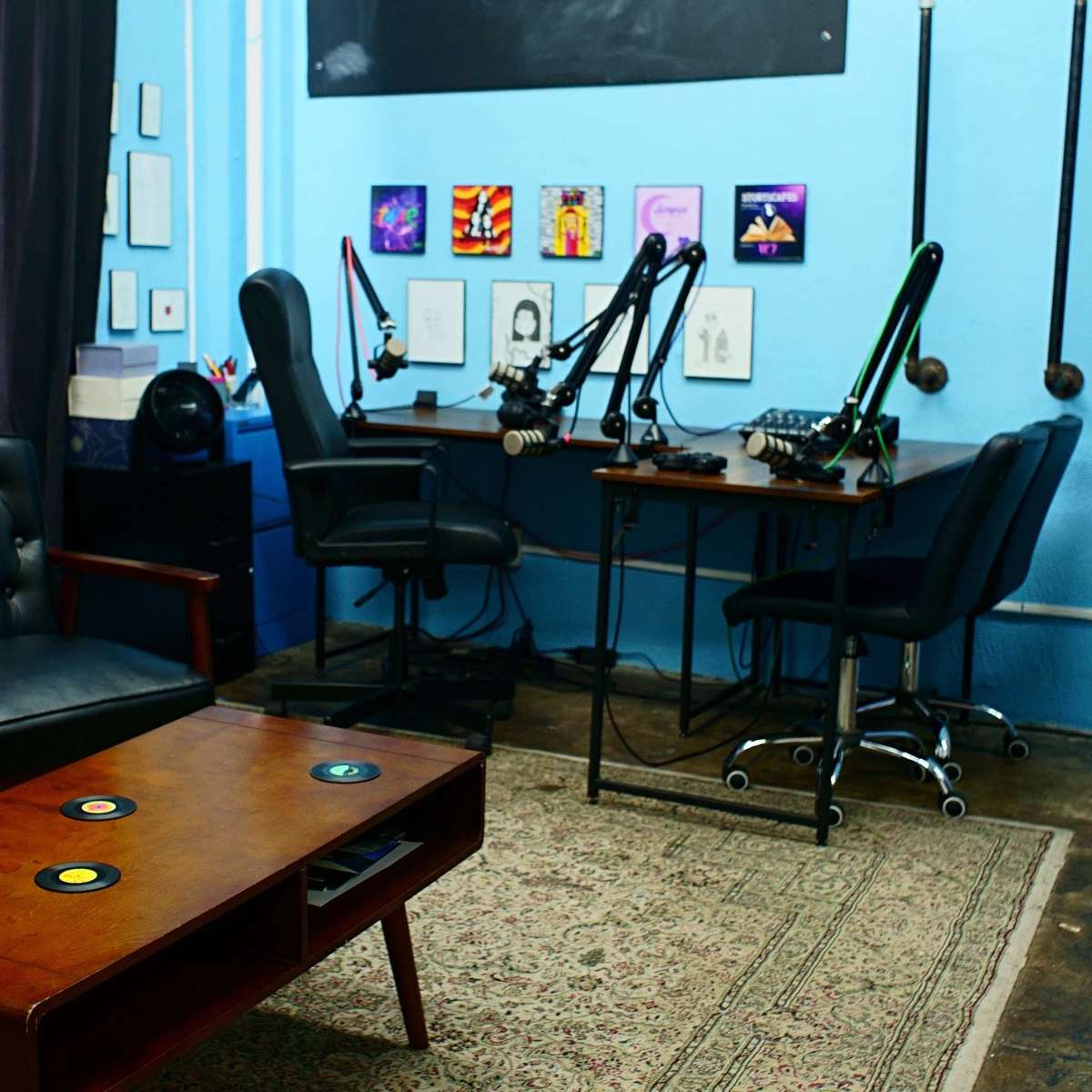 A room with a blue wall features a dark sofa, a wooden coffee table with records, and two workstations equipped with microphones and chairs.