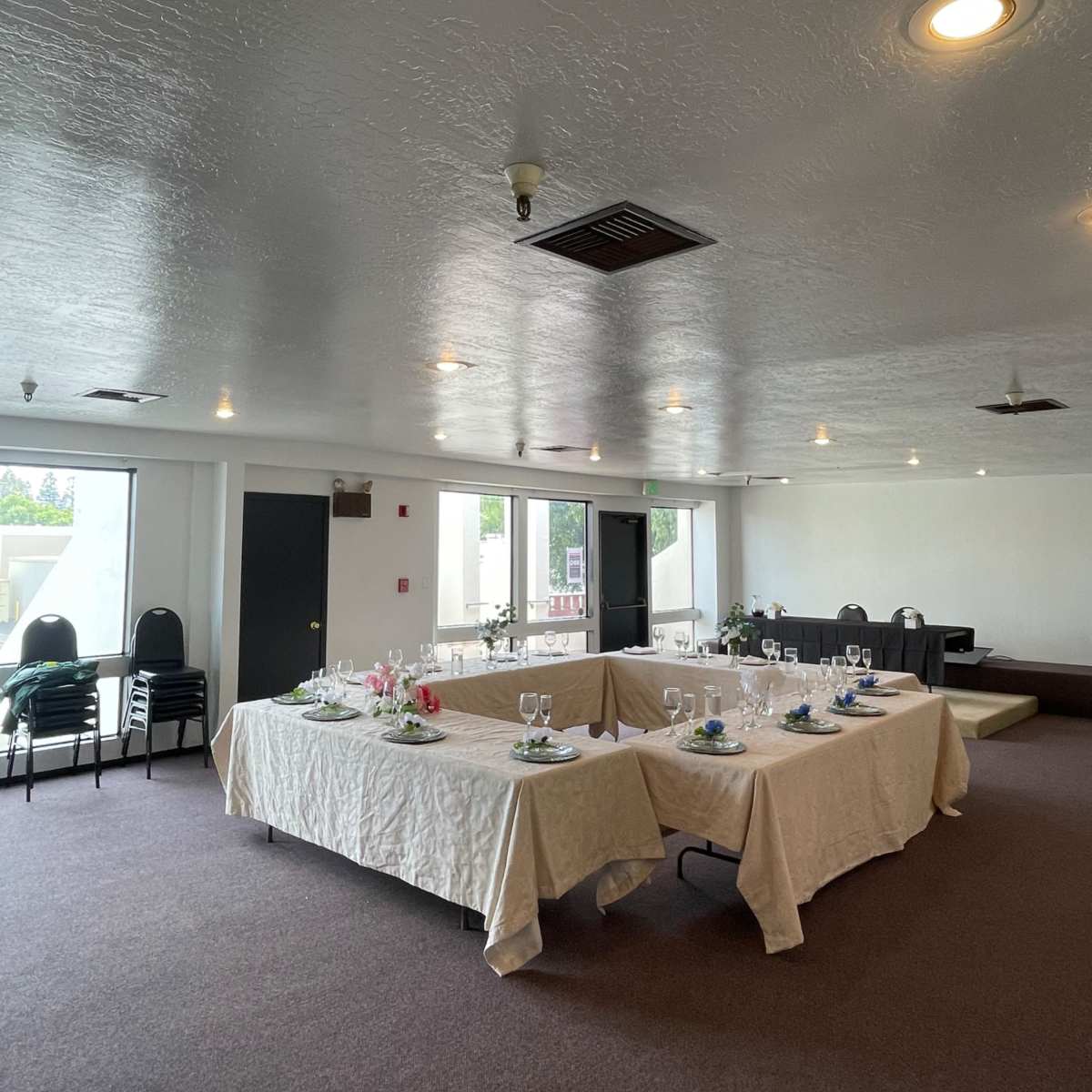 A spacious room features two tables set for a meal, surrounded by empty chairs and bright windows.