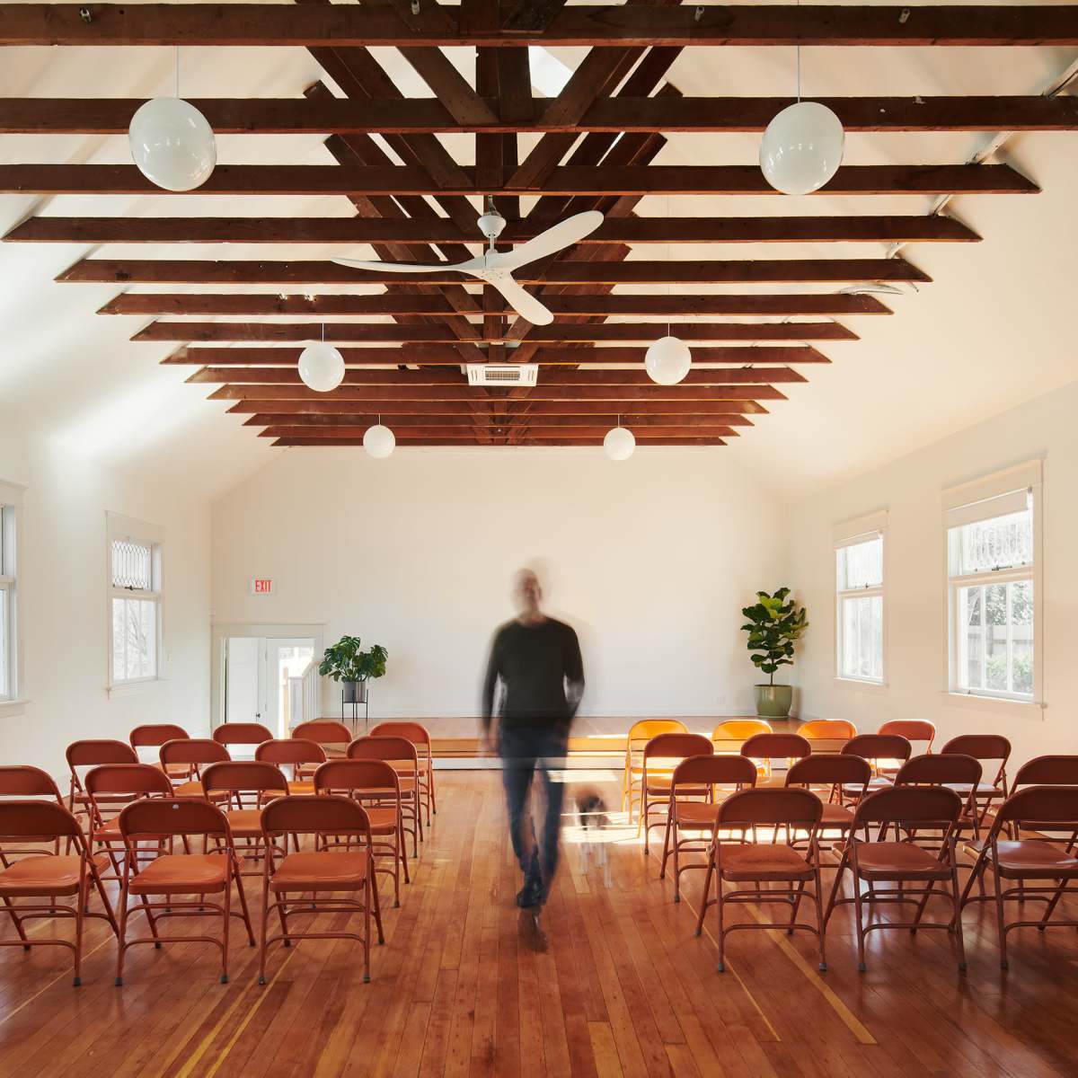 A man walks through a spacious, empty room with wooden beams, arranged chairs, and a potted plant.
