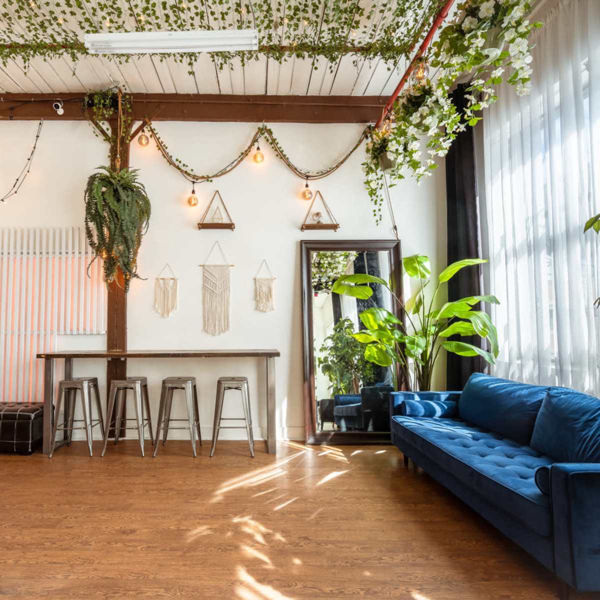 A modern, well-lit interior featuring a blue velvet sofa, tall indoor plants, and a bar-style table with stools against a backdrop of decorative greenery and hanging lights.