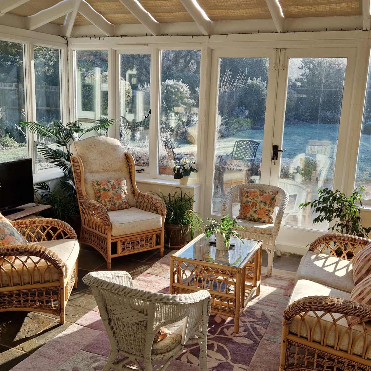 A sunlit conservatory features rattan furniture, including armchairs and a coffee table surrounded by indoor plants.