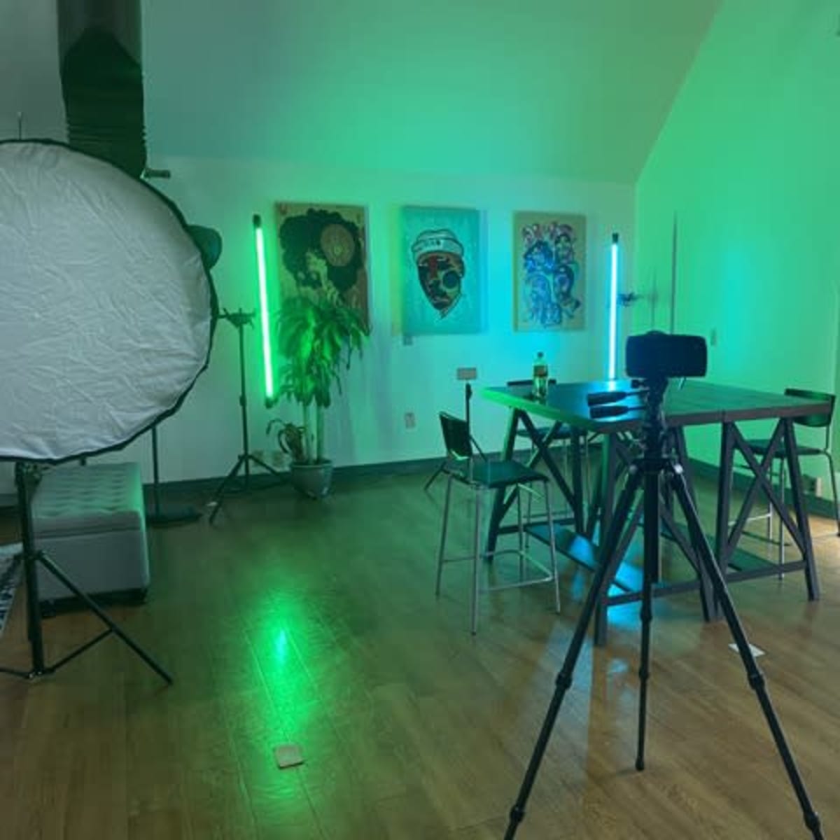 A camera tripod is set up in a well-lit room featuring a table, chairs, and artwork on the walls, with green LED lights and a large reflective disc nearby.
