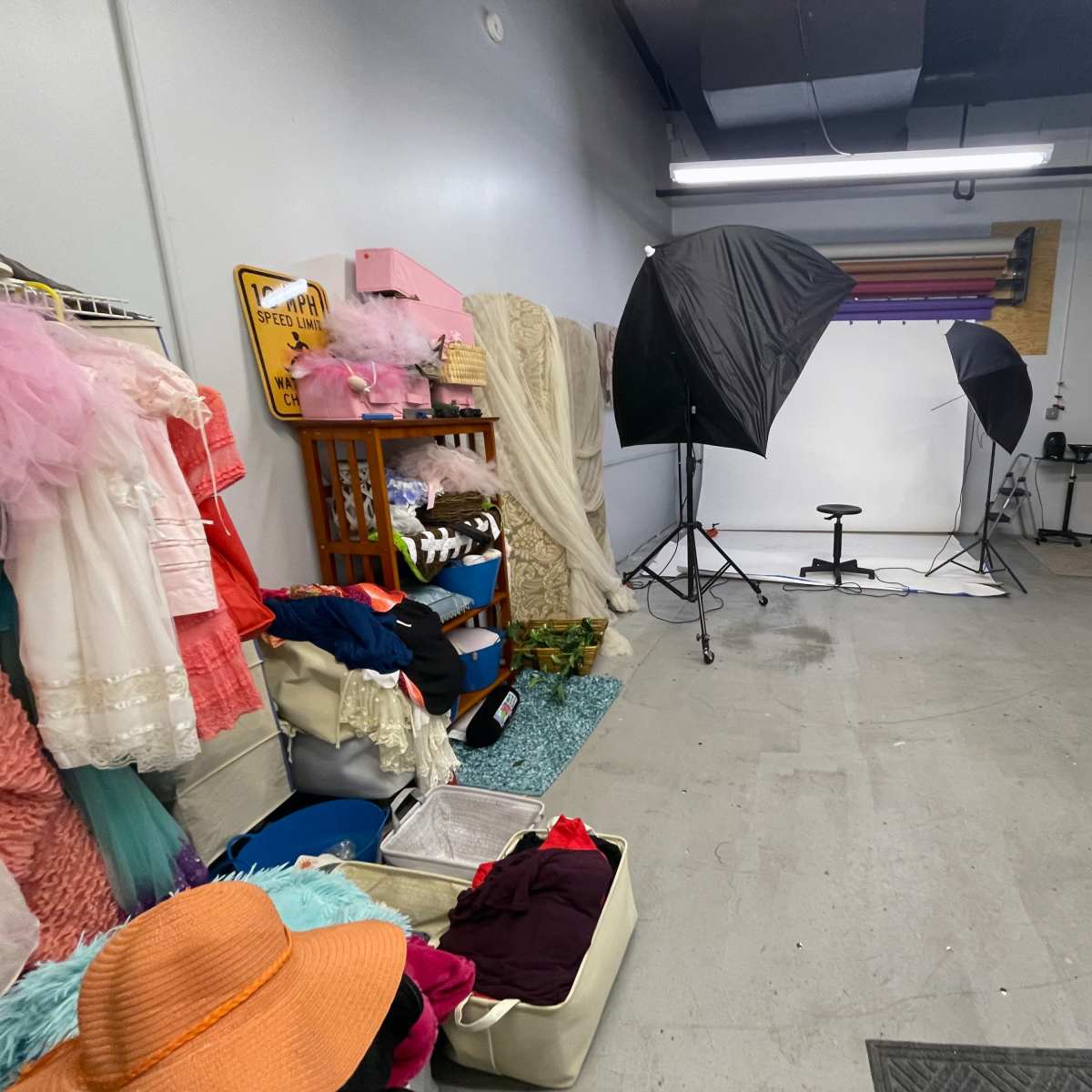A studio space with a backdrop setup for photography, flanked by various costumes and props stored in bins and on racks.