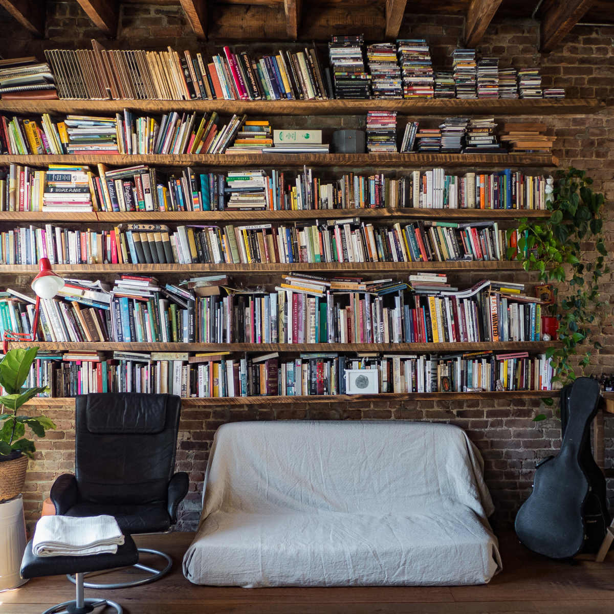 A cozy room features a wall of bookshelves filled with numerous books, a chair and a sofa covered with a white blanket, and a guitar leaning against the wall.