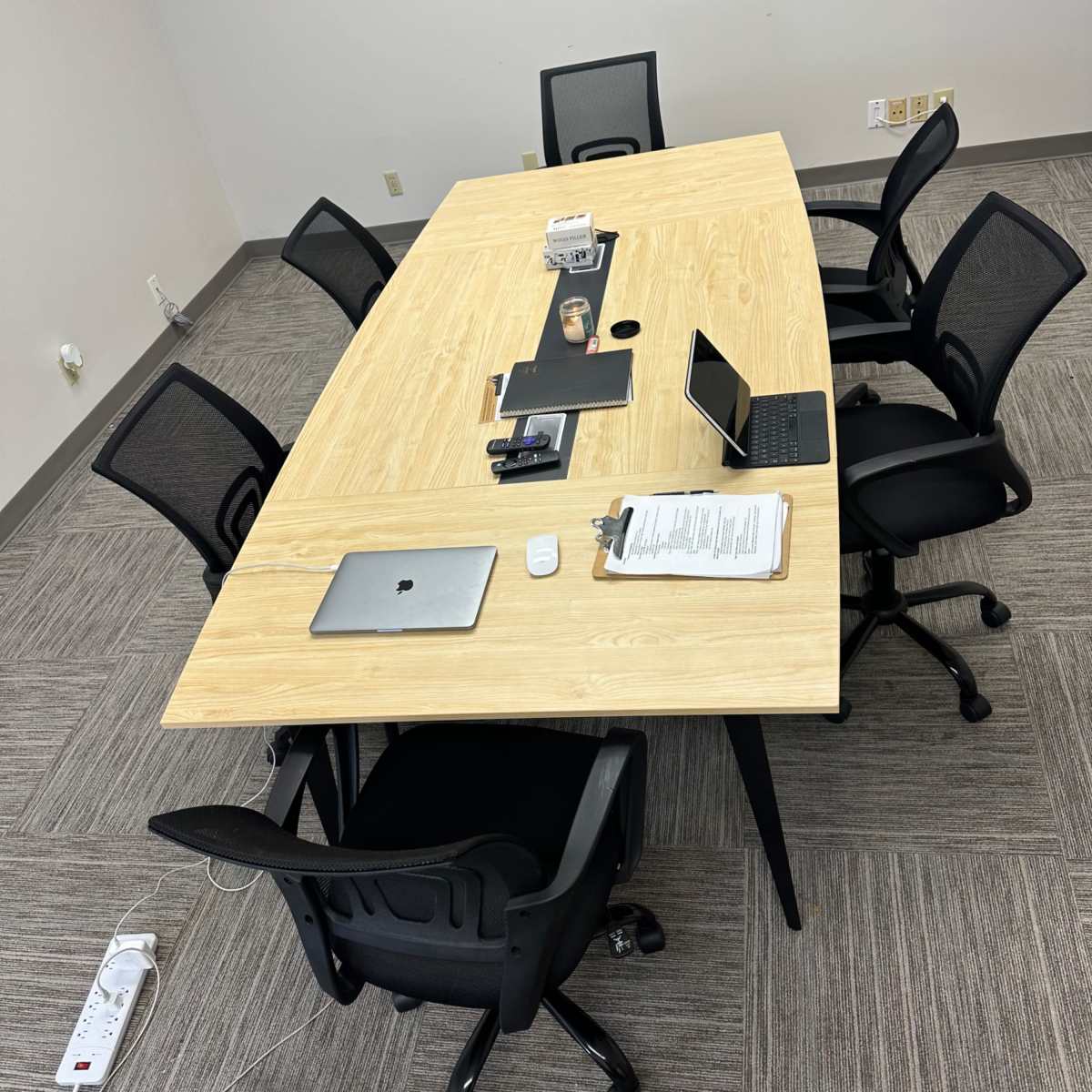 A large conference table with chairs surrounds it, and various office items including a laptop, tablet, and documents are placed on the table in a modern meeting room.
