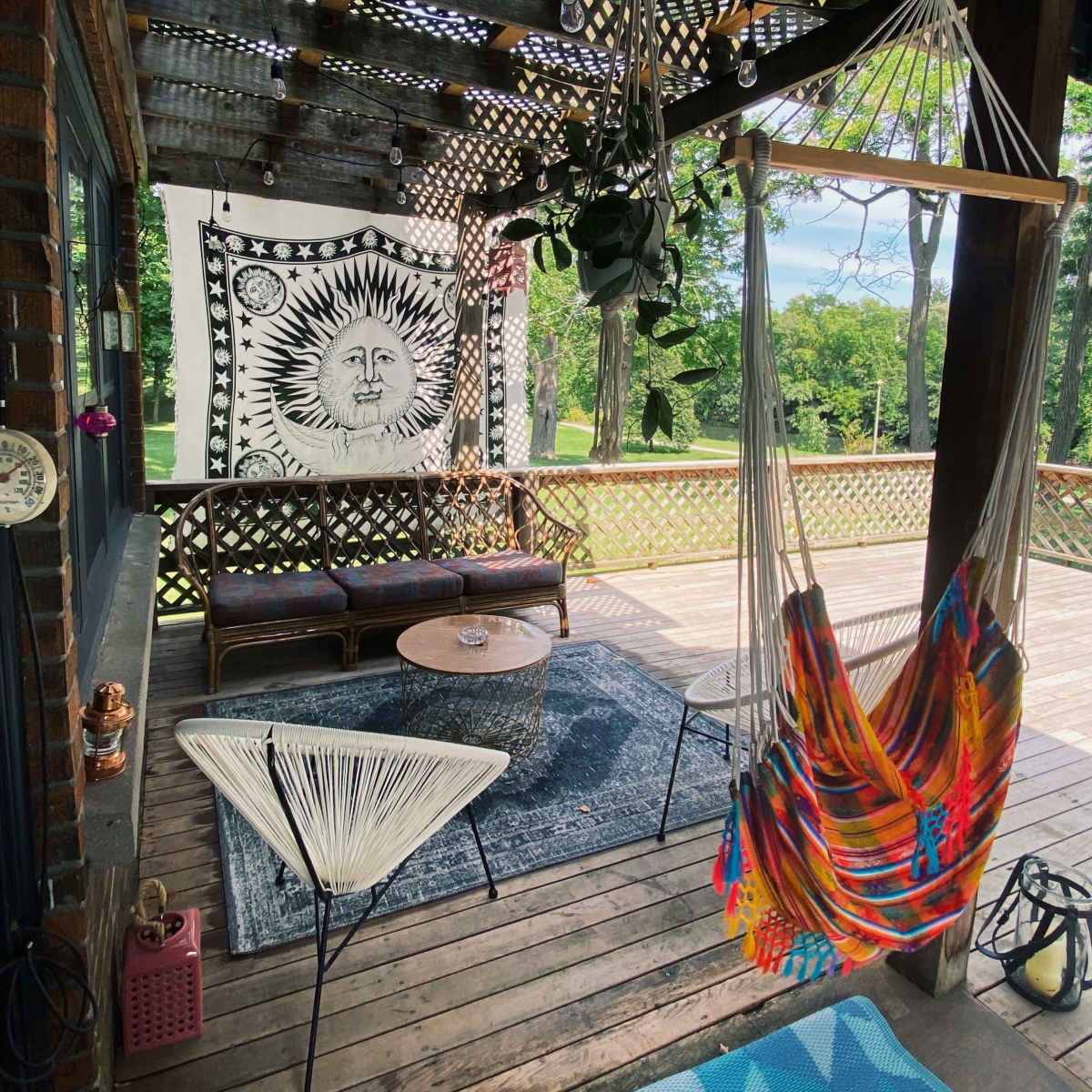 A covered porch with a sun-themed tapestry, colorful hammocks, and seating arrangements, surrounded by trees.