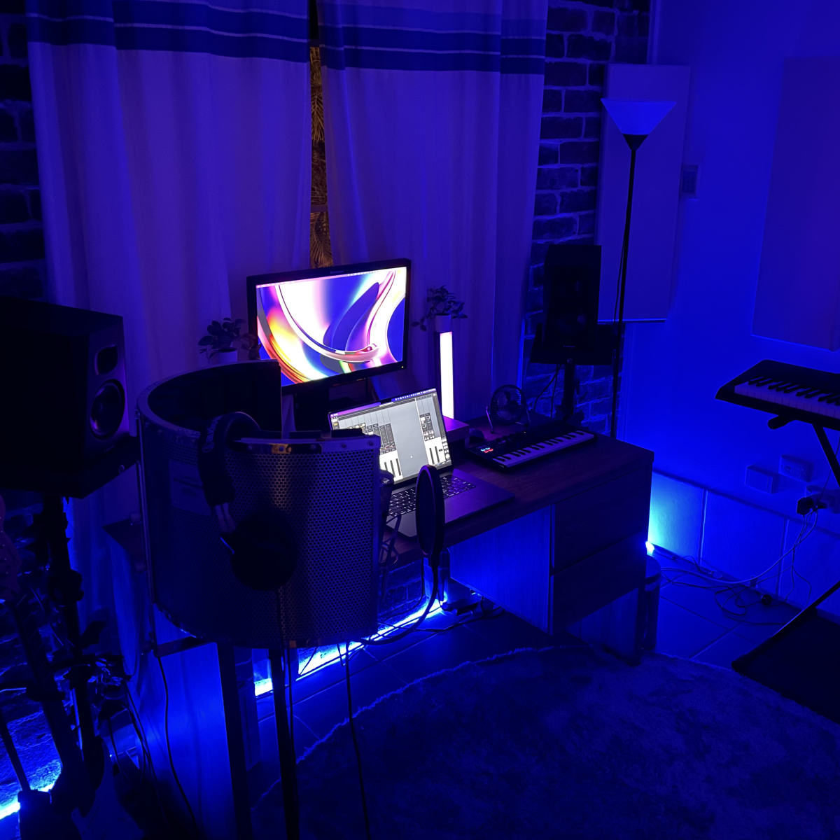 A music production studio with a desk, computer monitor, keyboard, speakers, and ambient lighting in blue tones.