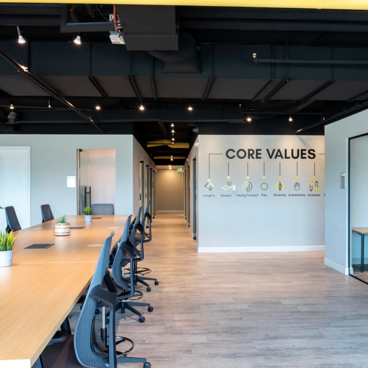 A modern office space featuring a long wooden table with black chairs, a wall displaying "CORE VALUES," and a glass-walled meeting room on the right.