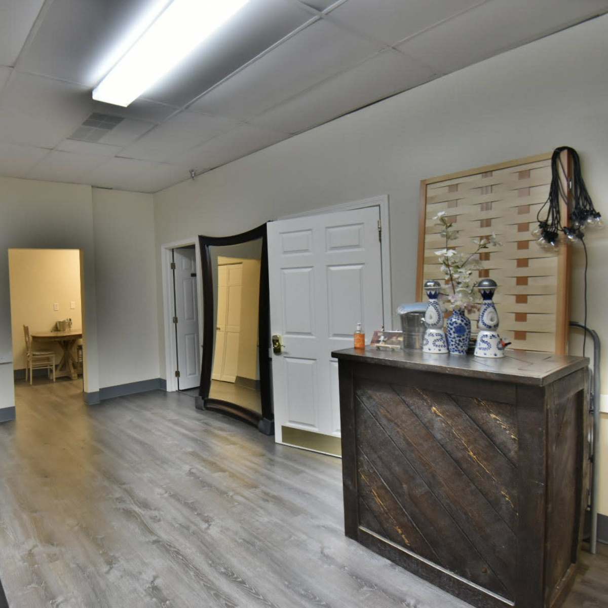 A spacious, well-lit room with a wooden counter, a large mirror, and a long wooden table along one wall, leading to a doorway at the back.