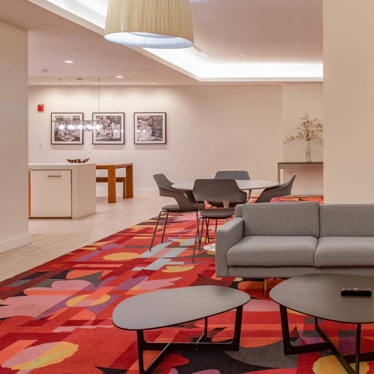 A modern lounge area with a gray couch, several small black tables, and a colorful rug, surrounded by seating and decorative wall art.