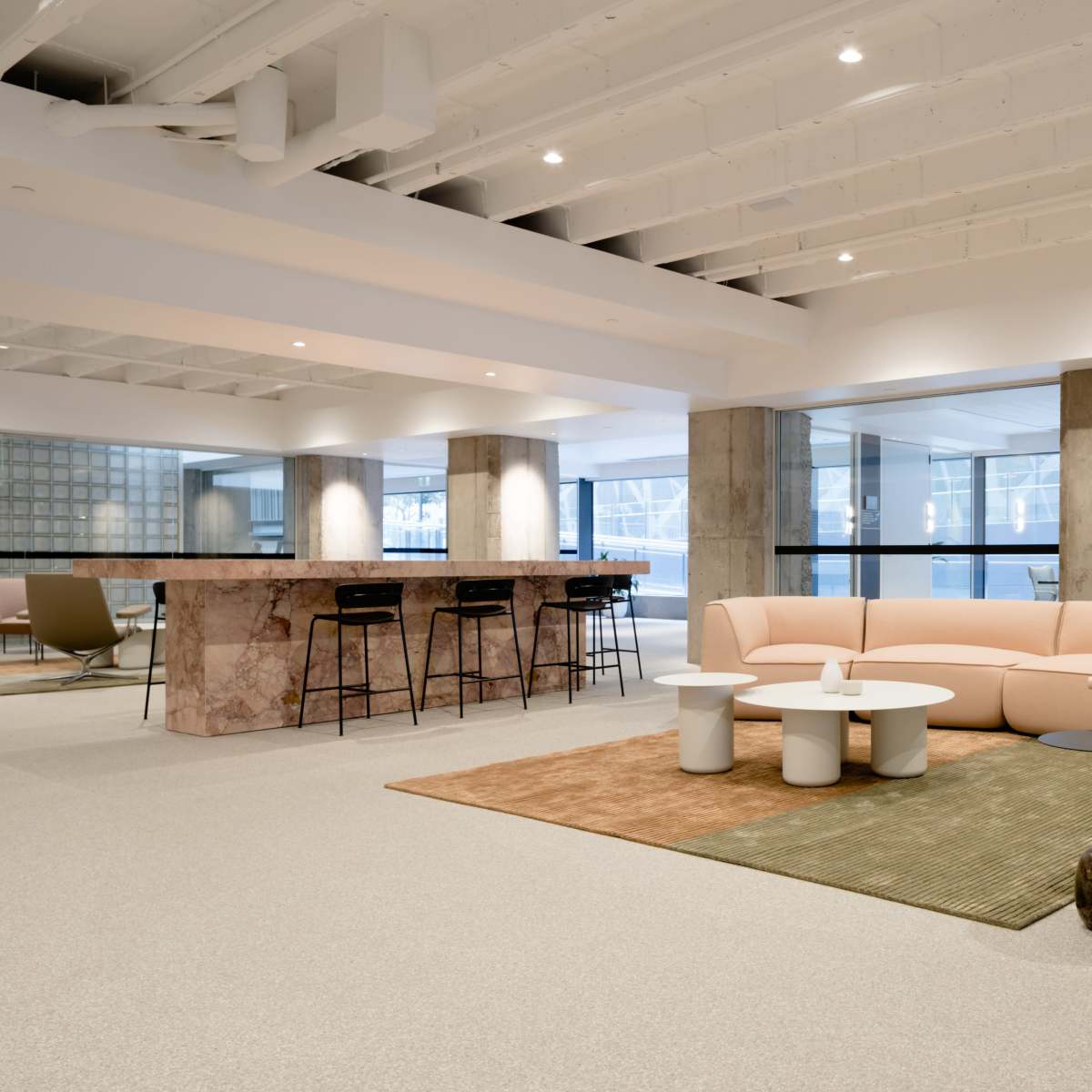 A modern office lounge area with a pink sofa, white tables, a marble bar, and large windows allowing natural light to enter.