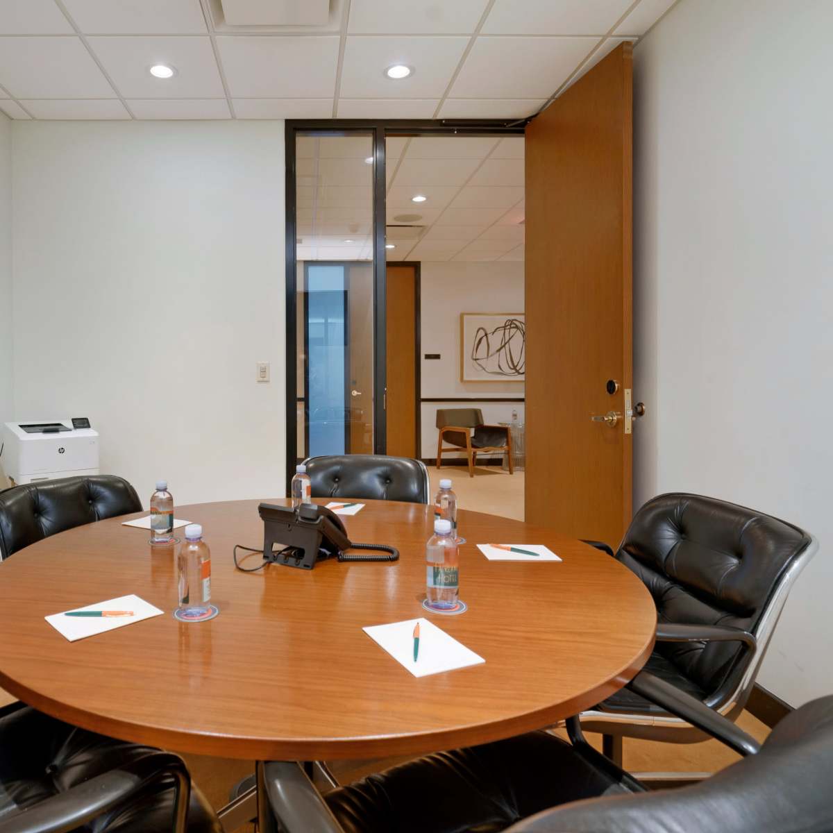 A round wooden table with black chairs and a phone is set up in a conference room, featuring water bottles and notepads at each seat.
