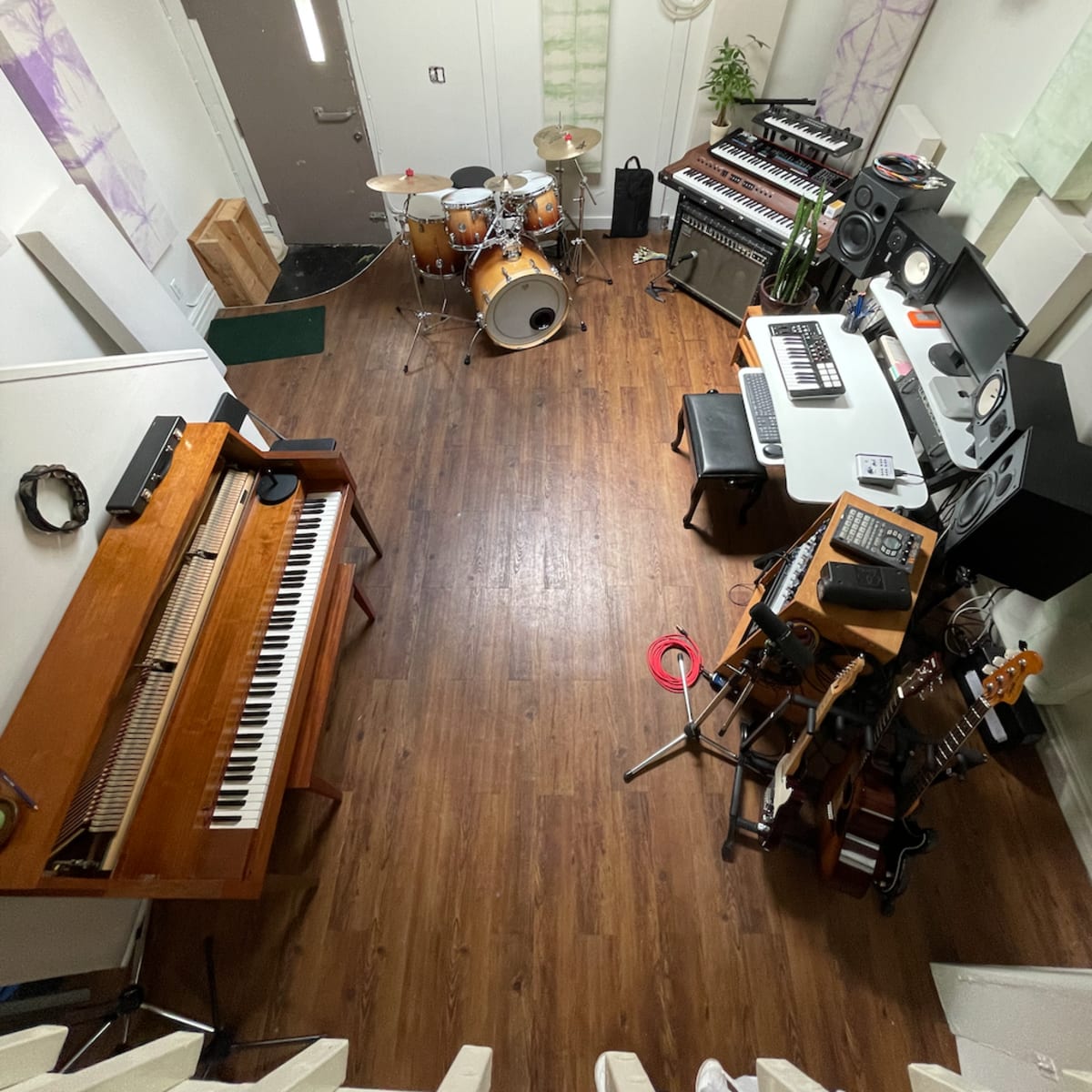 A music studio featuring a drum set, a keyboard, synthesizers, and various guitars arranged in a wooden-floored room.