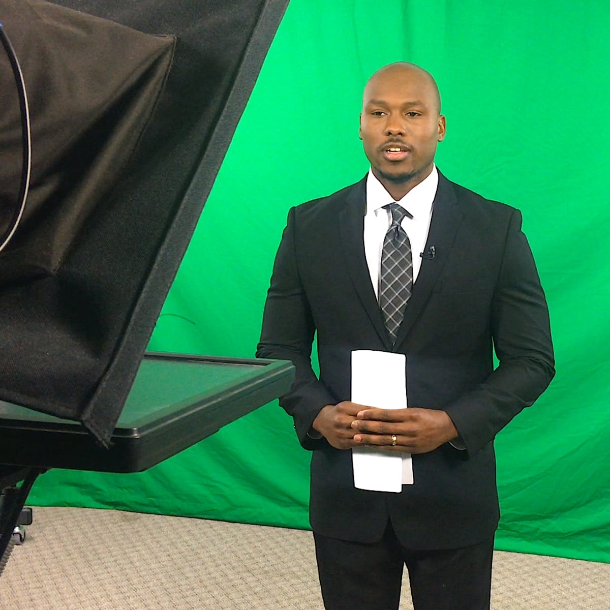 A man in a suit stands in front of a green screen, holding a stack of papers and facing the camera.