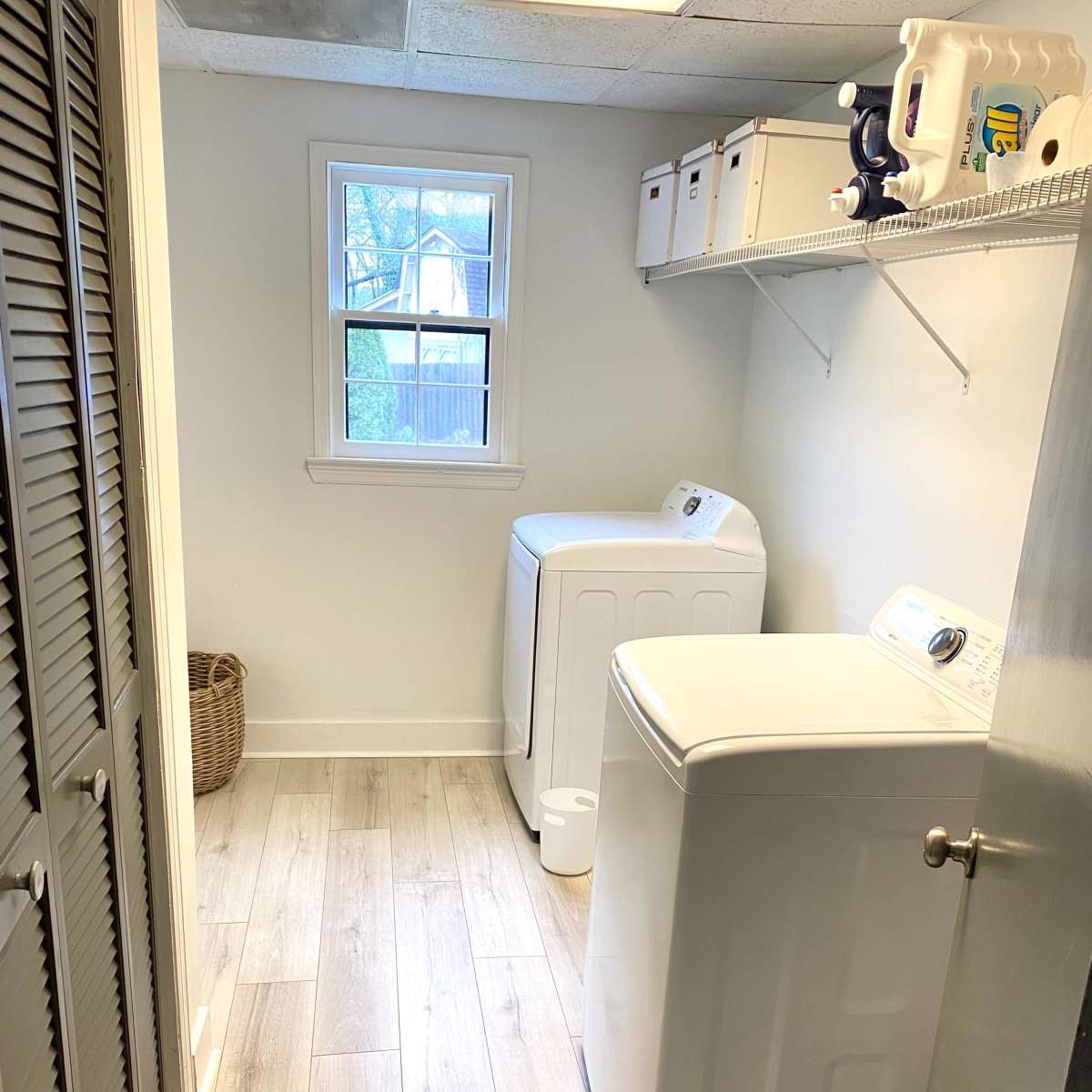 A laundry room featuring two white washing machines, a window, and neatly organized storage boxes on a shelf.
