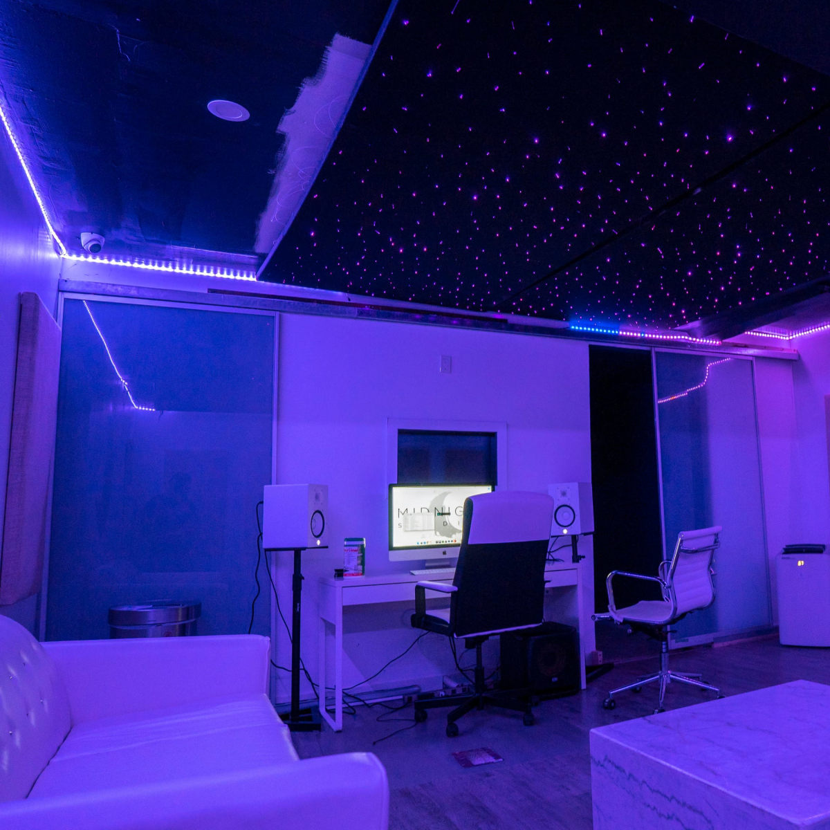 A modern recording studio with purple lighting, a white sofa, a desk with studio monitors, and a screen in the background.