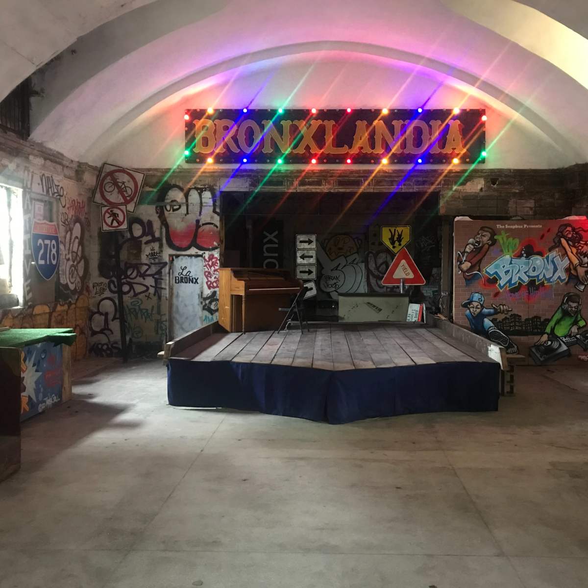 An underground room with colorful graffiti on the walls, a wooden stage in the center, and lights spelling "BRONXLAND" above.