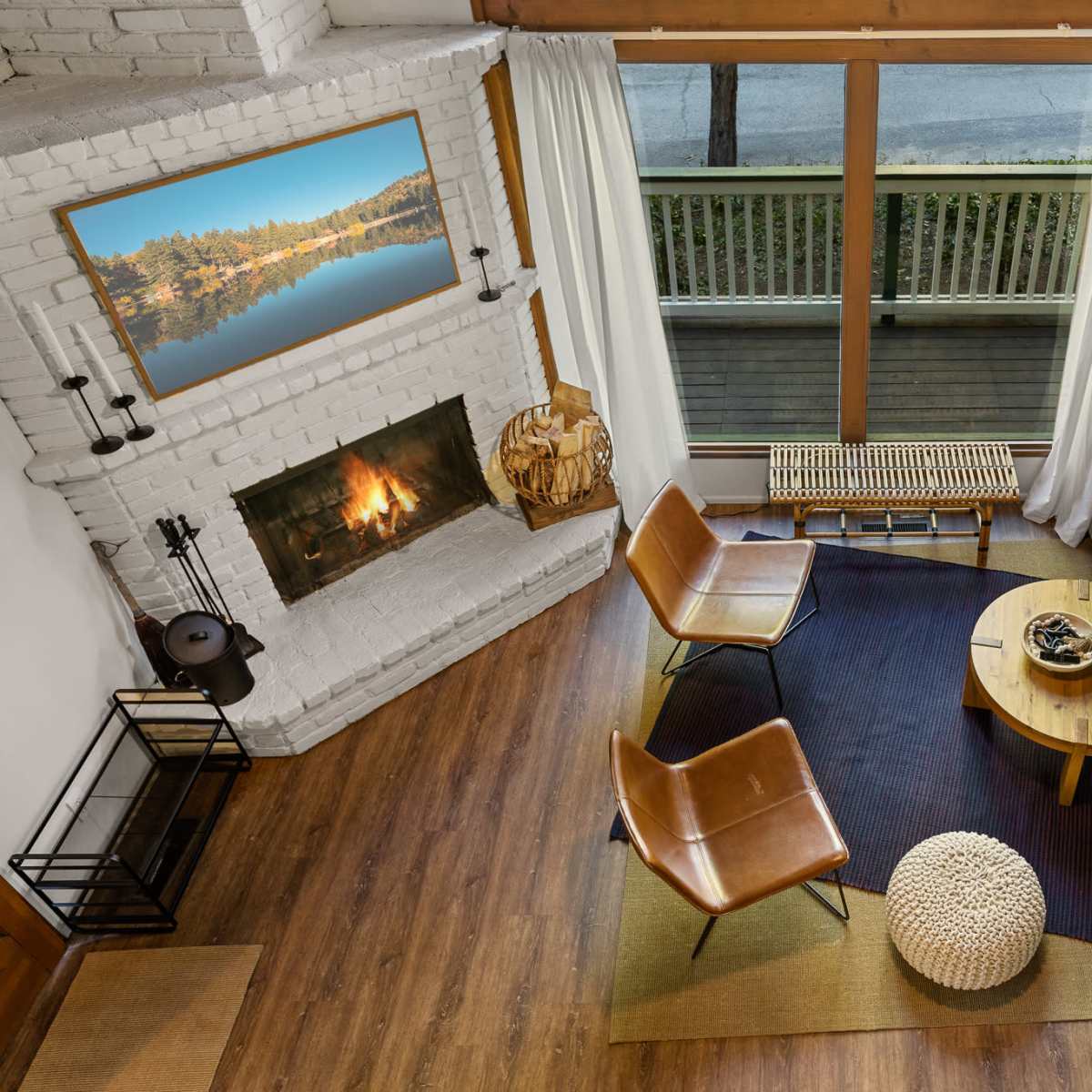 A cozy living room with a white-bricked fireplace, a large sectional sofa, two leather chairs, a round coffee table, and a view of a deck through large glass doors.