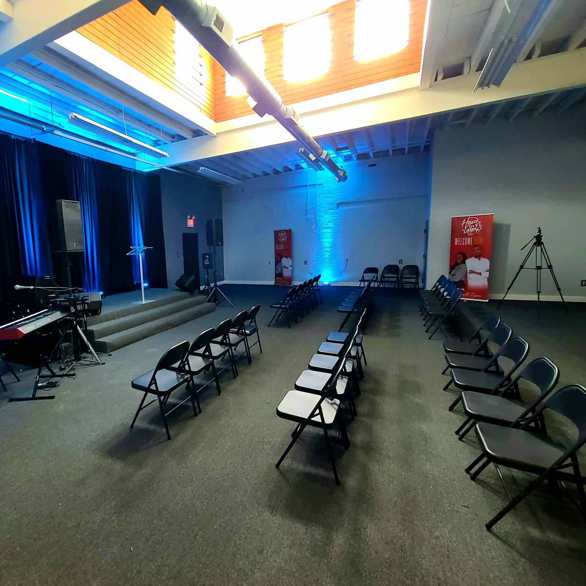 A room set up for an event, featuring rows of black chairs facing a stage with musical instruments and blue lighting.