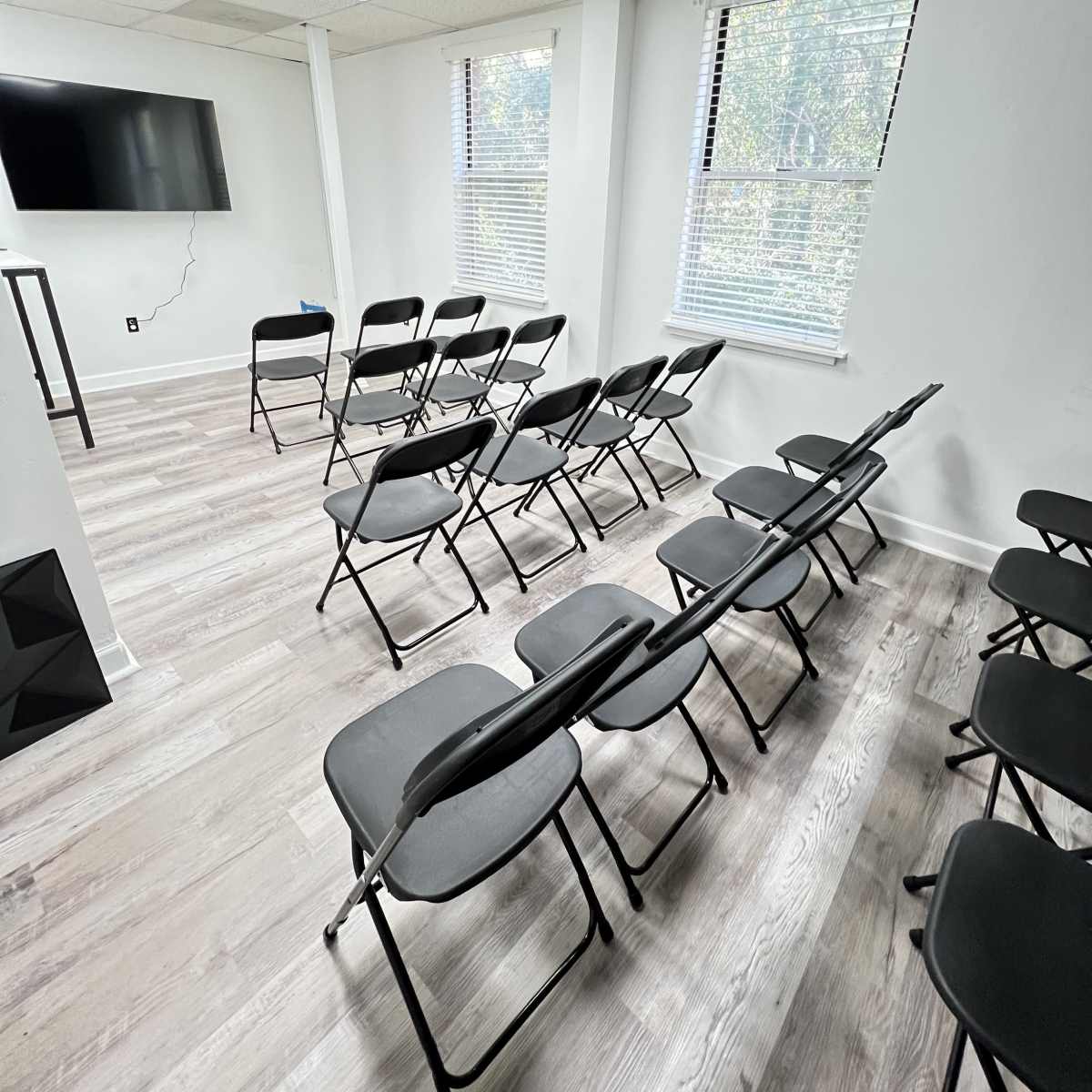 A small, bright room arranged with several black folding chairs set in rows, facing a wall-mounted television.