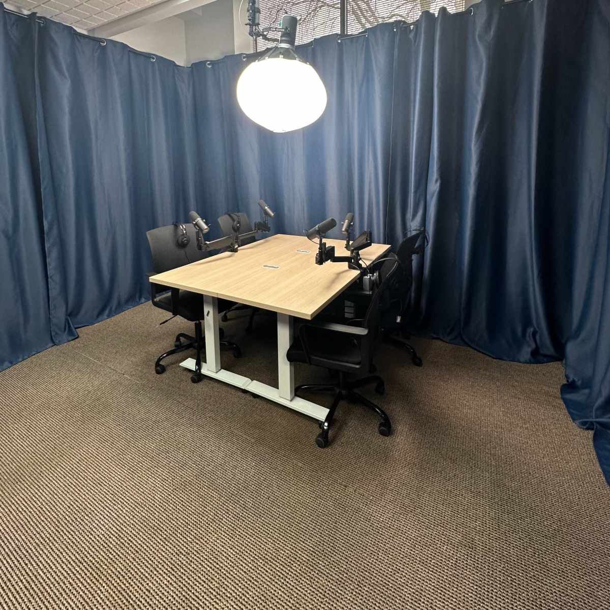 A small meeting room features a central wooden table surrounded by four black chairs and is illuminated by a large overhead light, with blue curtains creating a private space.