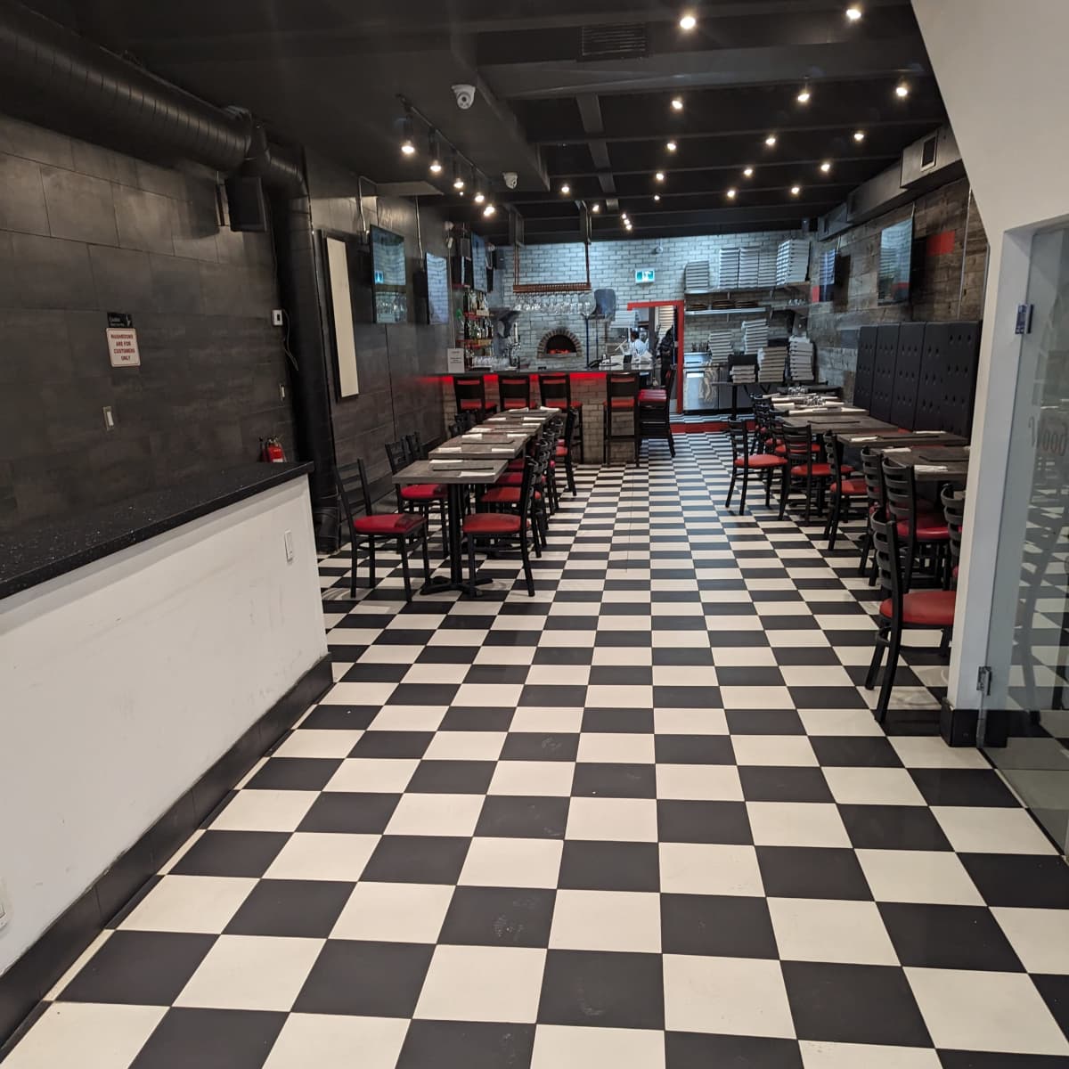 The interior of a restaurant features a black and white checkered floor with tables and chairs arranged in rows, leading to a bar area at the back.