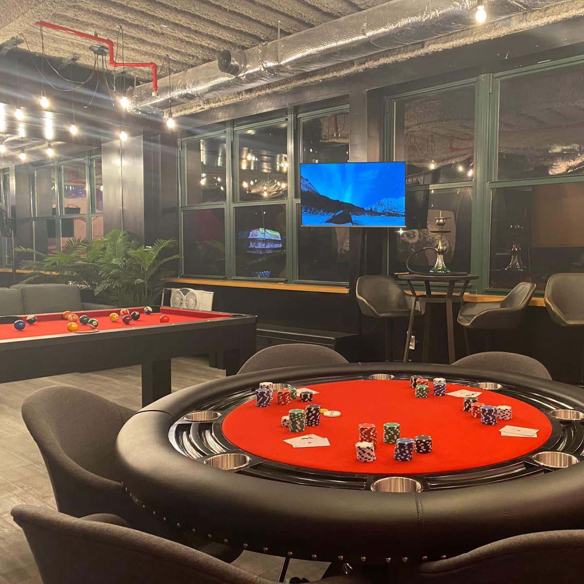 A poker table with chips arranged on a red felt surface, surrounded by modern seating and a television displaying a scenic image in a well-lit room.