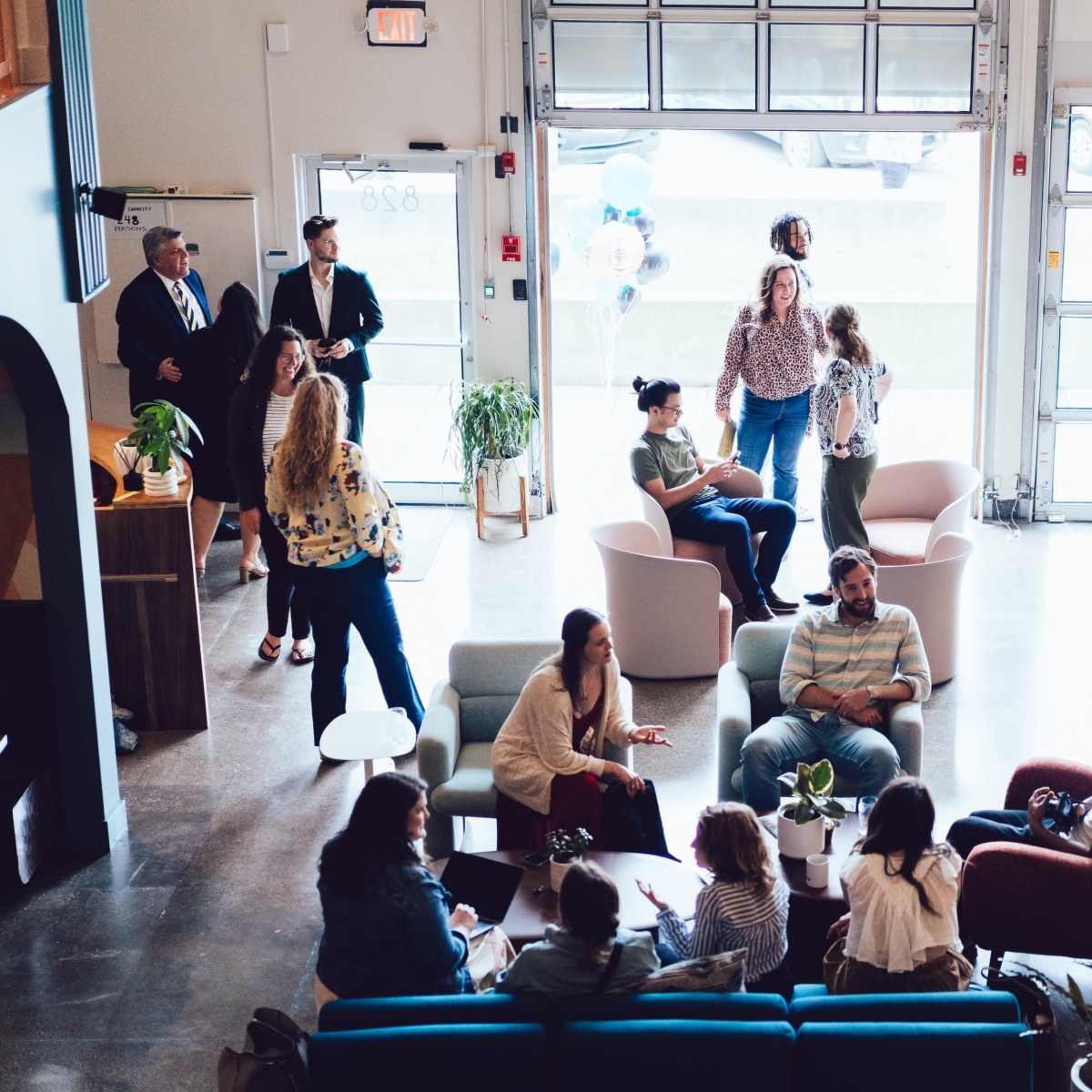 A spacious indoor gathering area with various groups of people engaged in conversation, seated in modern chairs and sofas, while large garage doors are open to the outside.