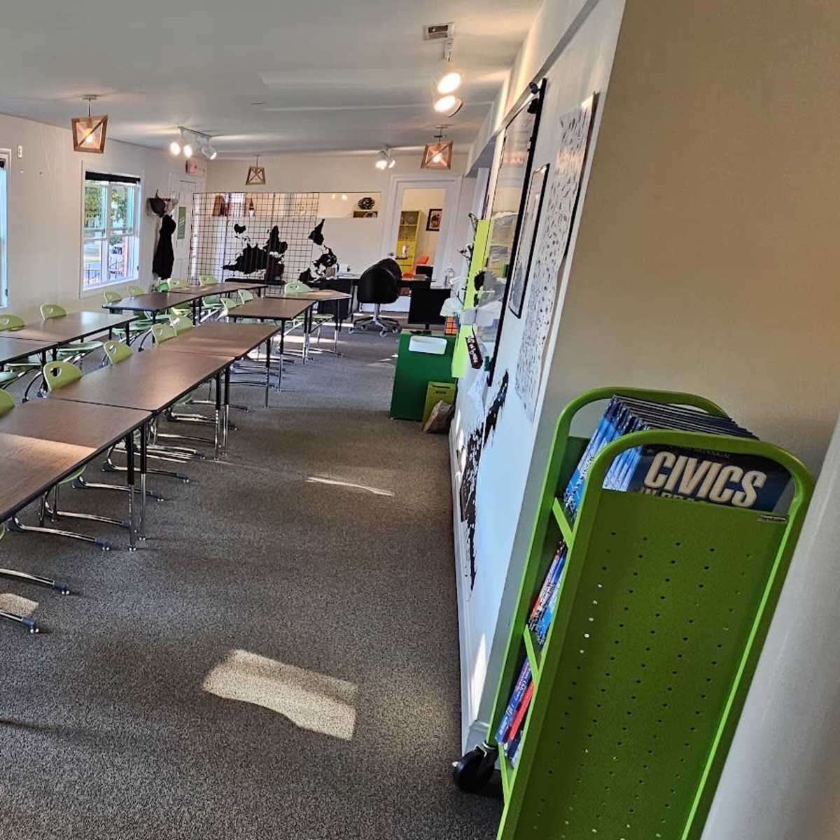 A classroom features multiple rows of tables and green chairs arranged on a carpeted floor, with large windows allowing natural light.
