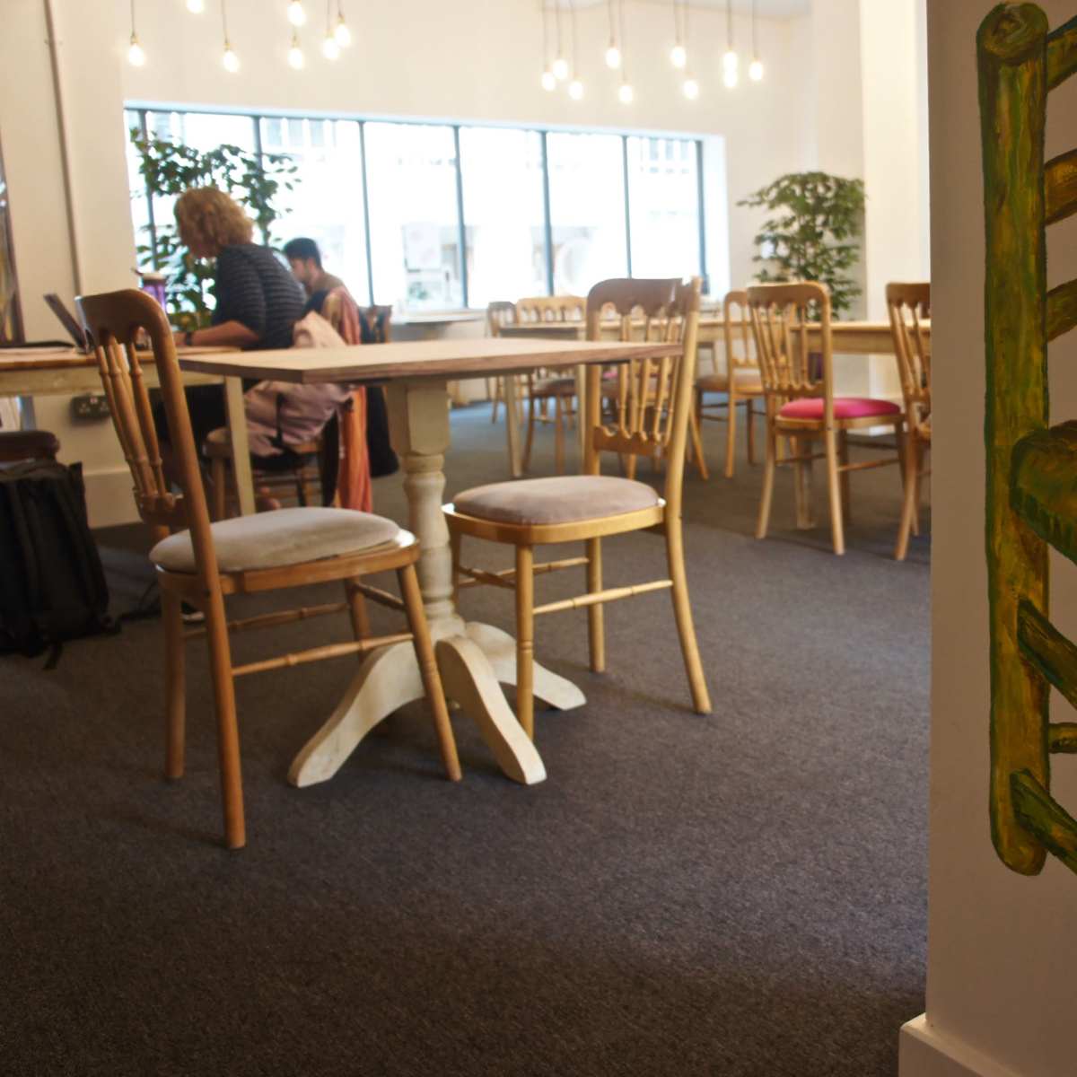 A spacious, modern café scene with wooden chairs, a large table, and a painted green chair on the wall.