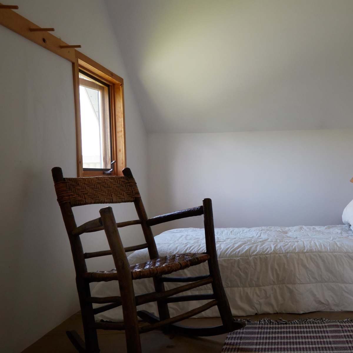A minimalist bedroom with a single bed, a rocking chair, and a lamp beside a window.