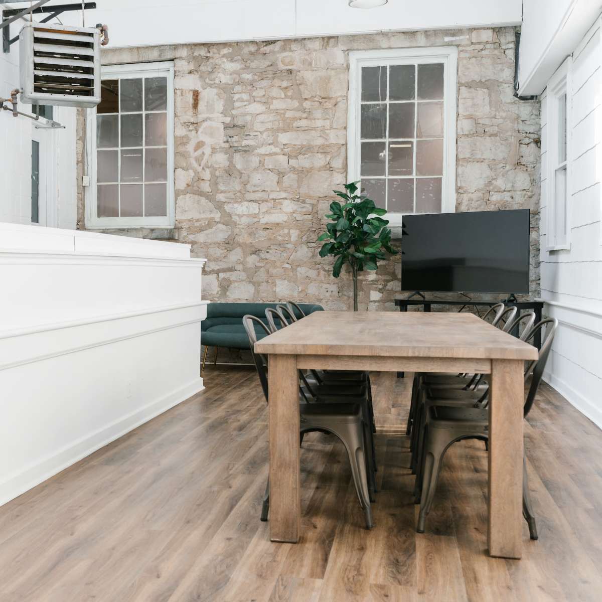 A spacious meeting room features a long wooden table surrounded by metal chairs, with large windows and a stone wall in the background.
