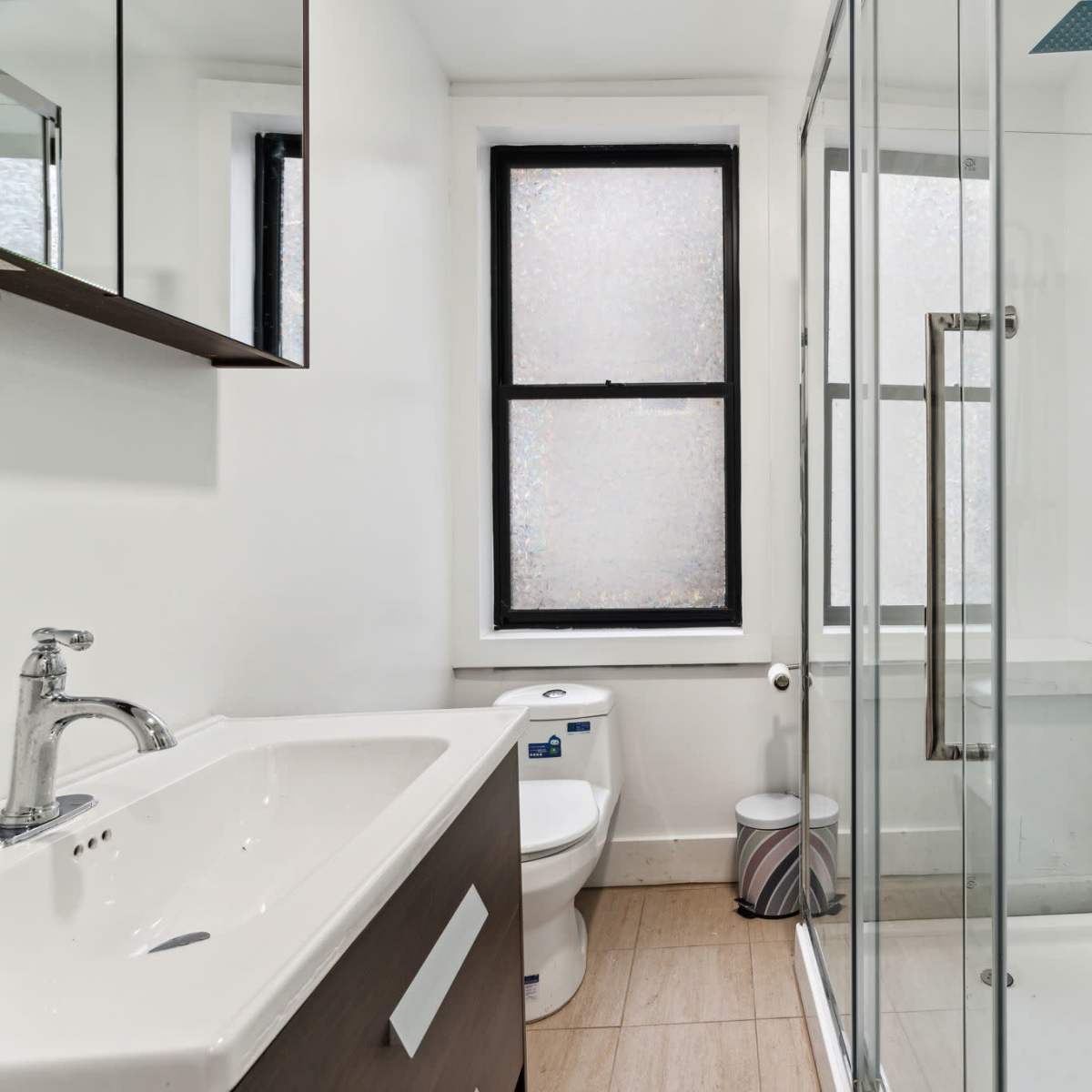 A modern bathroom with a glass shower enclosure, a toilet, a sink with a dark wood vanity, and large windows.