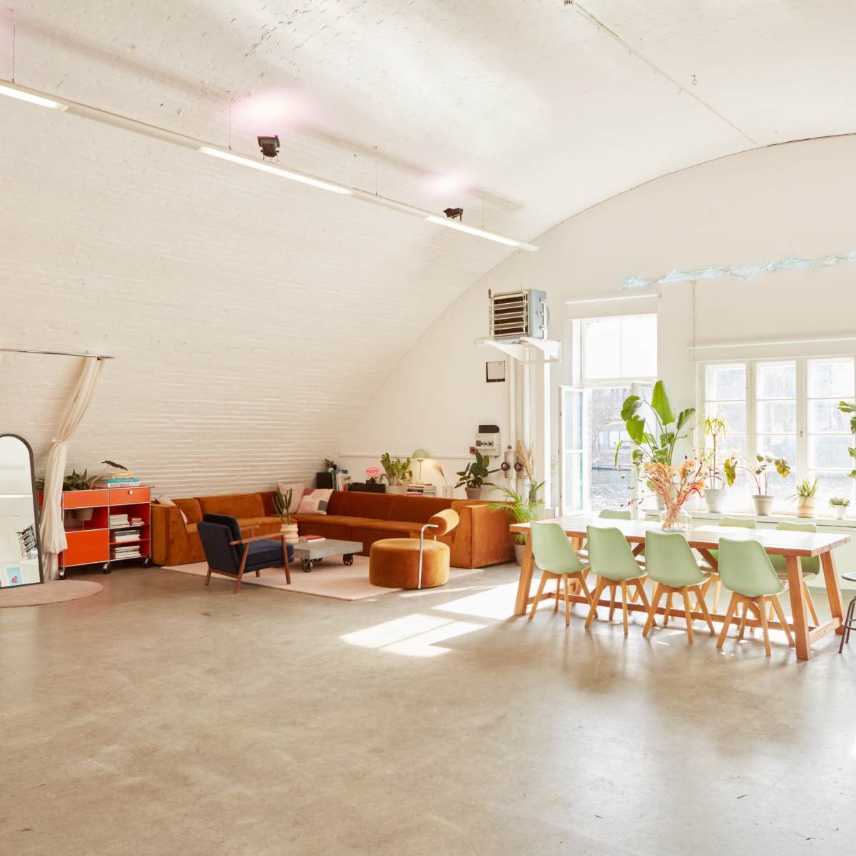 A spacious, well-lit room with a curved ceiling, featuring a seating area with a sofa, a dining table, and large windows filled with plants.