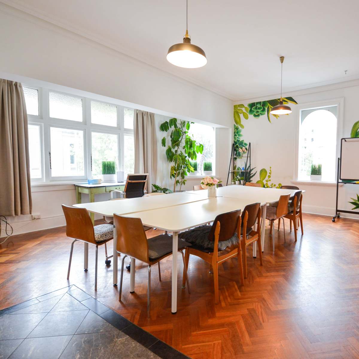 A spacious meeting room features a large table surrounded by chairs, with large windows and plants decorating the walls.