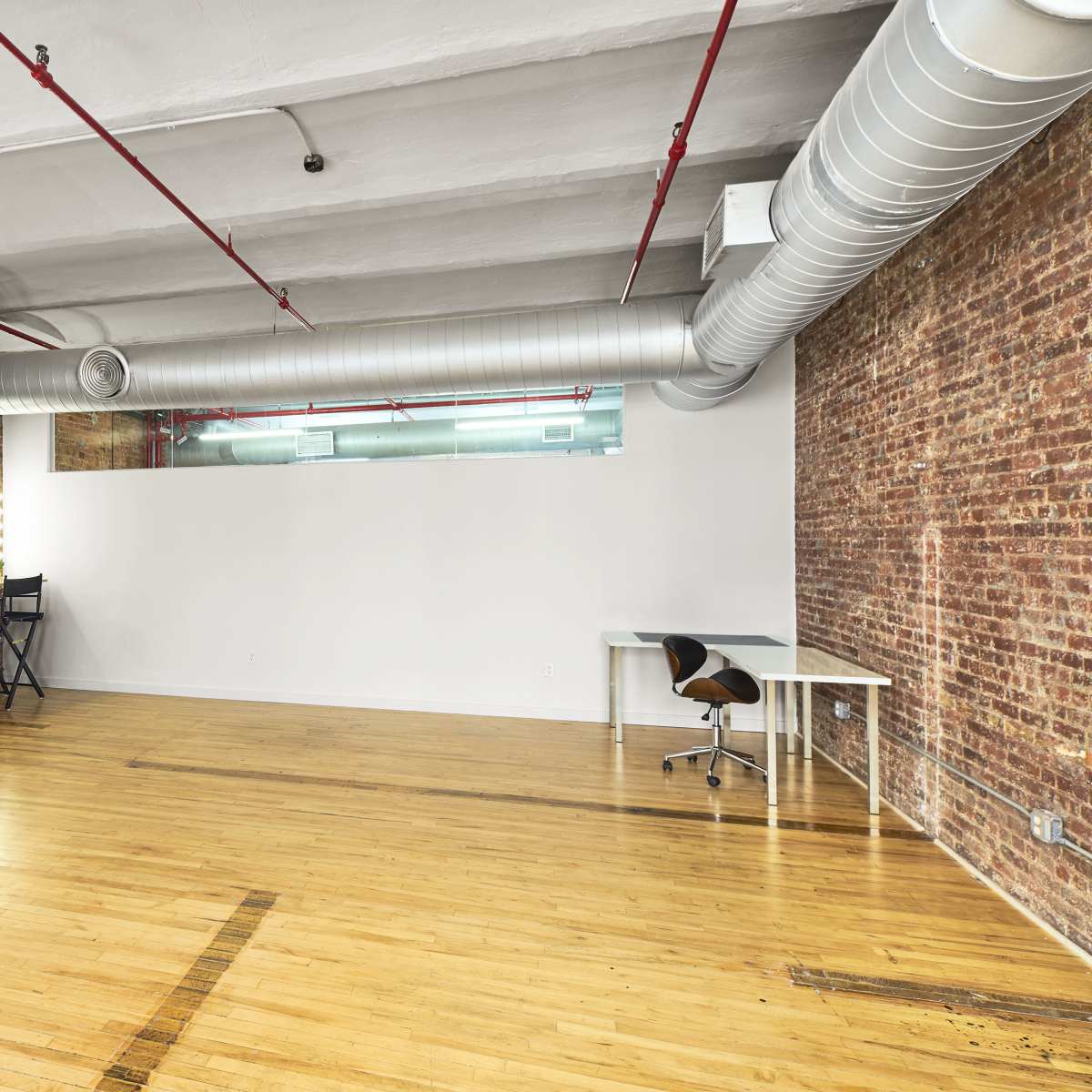 An empty room with wooden floors, exposed brick walls, and industrial-style ductwork overhead, featuring a vanity mirror on one side and a simple desk and chair on the other.