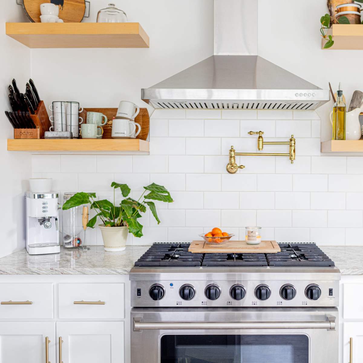 A modern kitchen with a stainless steel gas stove, a gold wall-mounted faucet, open shelving displaying dishes and plants, and a marble countertop.