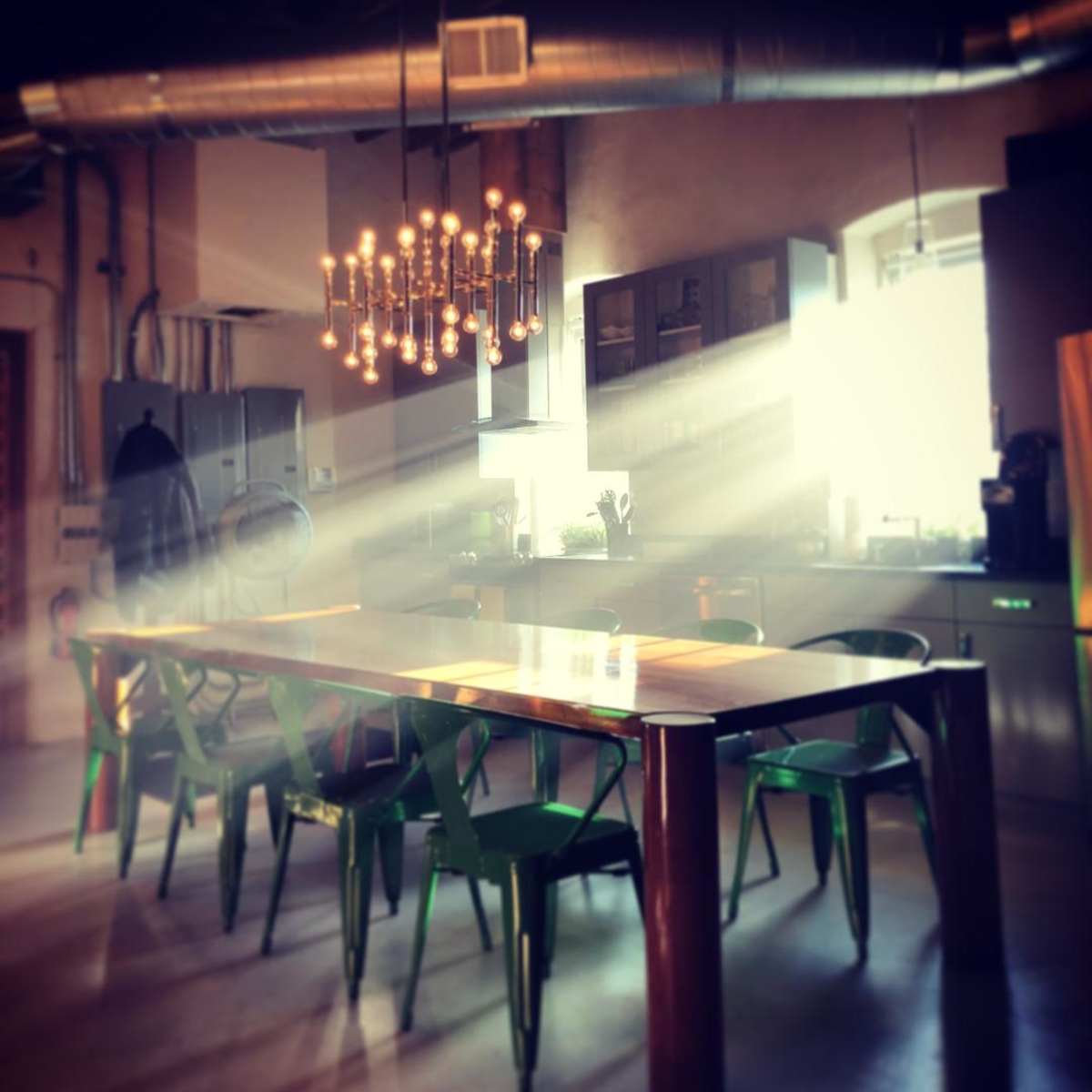 A large wooden dining table surrounded by green chairs is illuminated by sunlight streaming through a window in a modern kitchen.