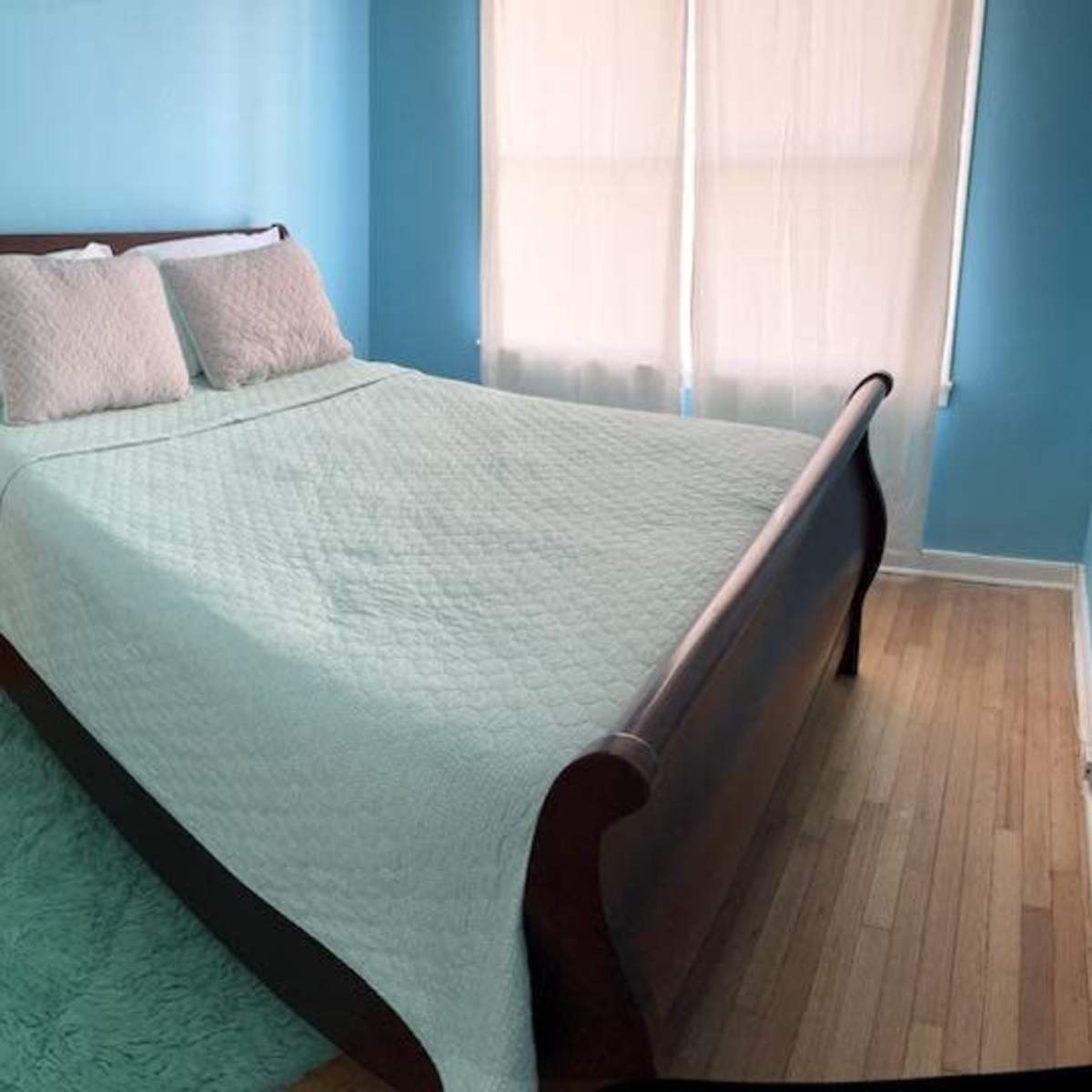 A bedroom with a wooden sleigh bed covered in a light quilt, a nightstand with a lamp, and large windows with sheer curtains on a hardwood floor.