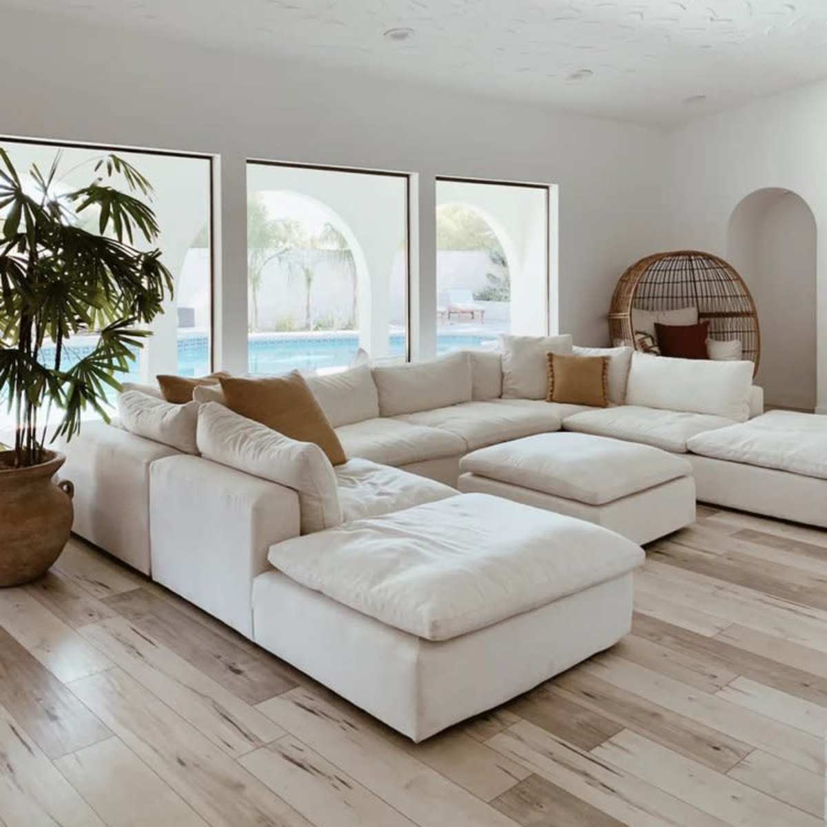 A light-filled living room with a large white sectional sofa, a decorative plant, and arched windows revealing a pool view.