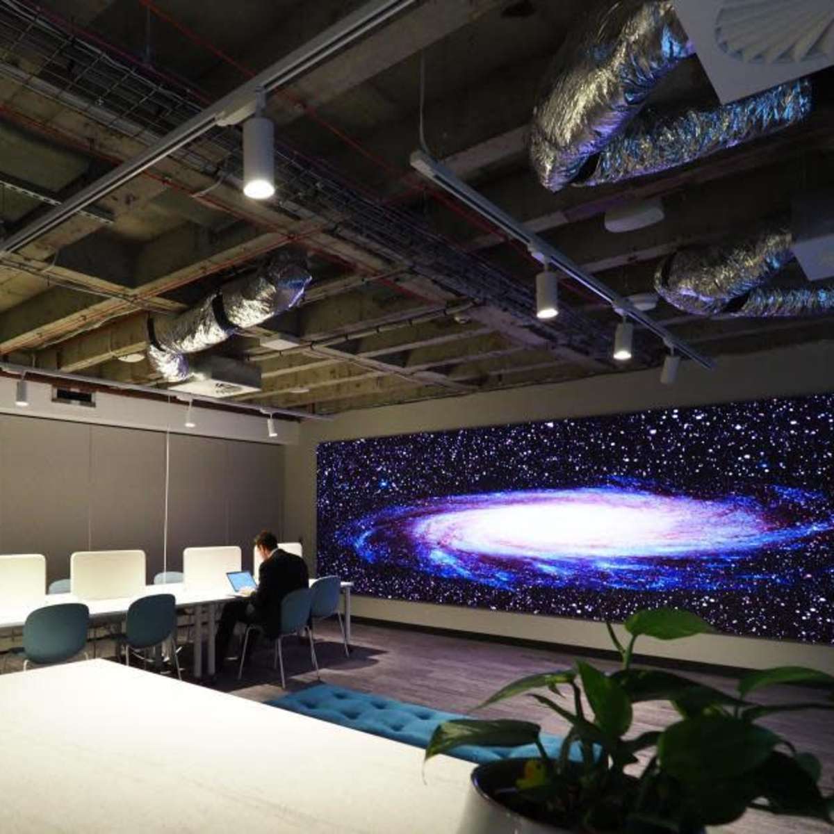 A modern office space with a large screen displaying a galaxy, seating areas, and industrial-style ceiling features.